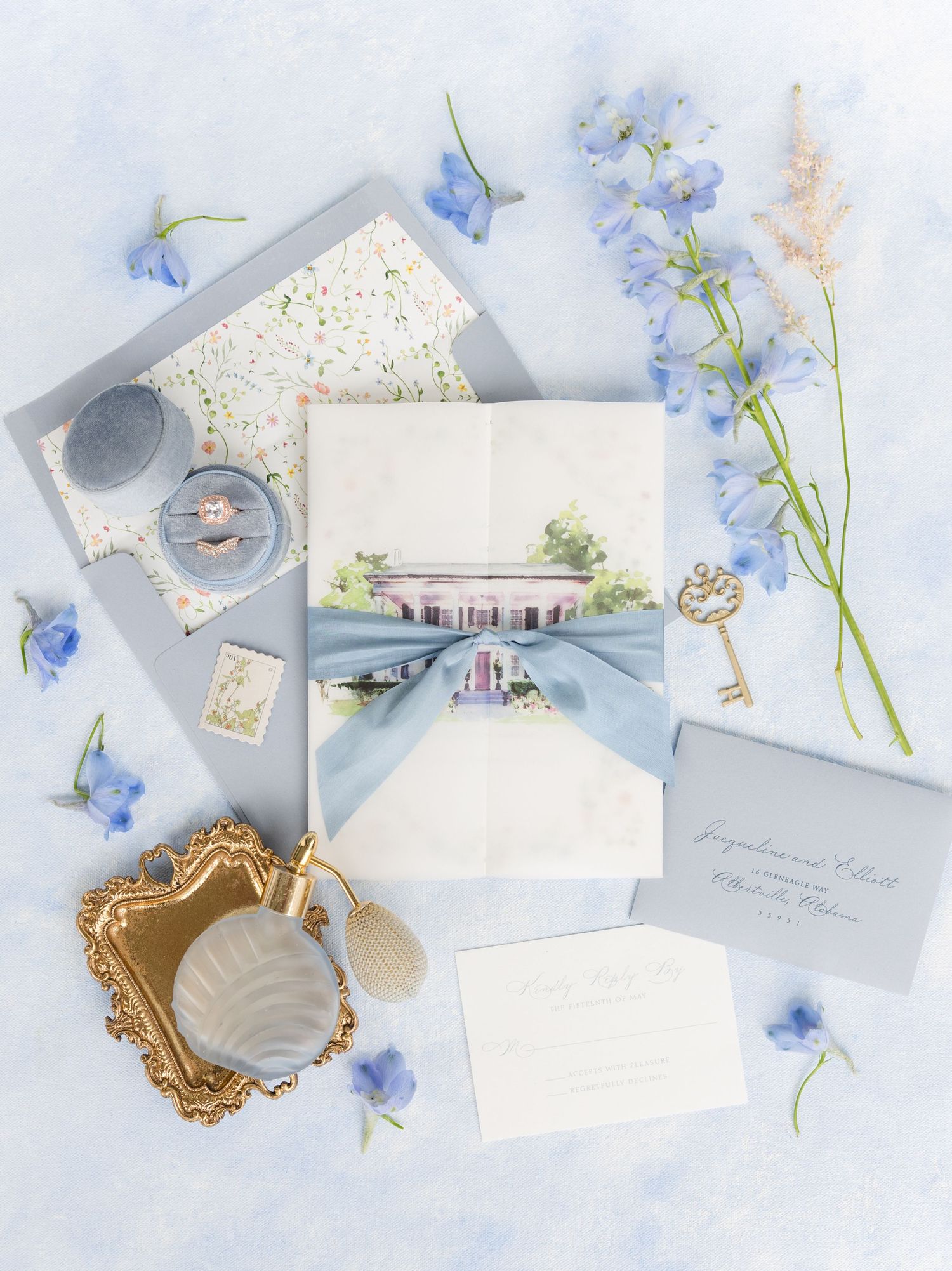 Elegant wedding invitation suite with dusty blue ribbon, floral watercolor venue illustration, ring, perfume bottle, and blue flowers.