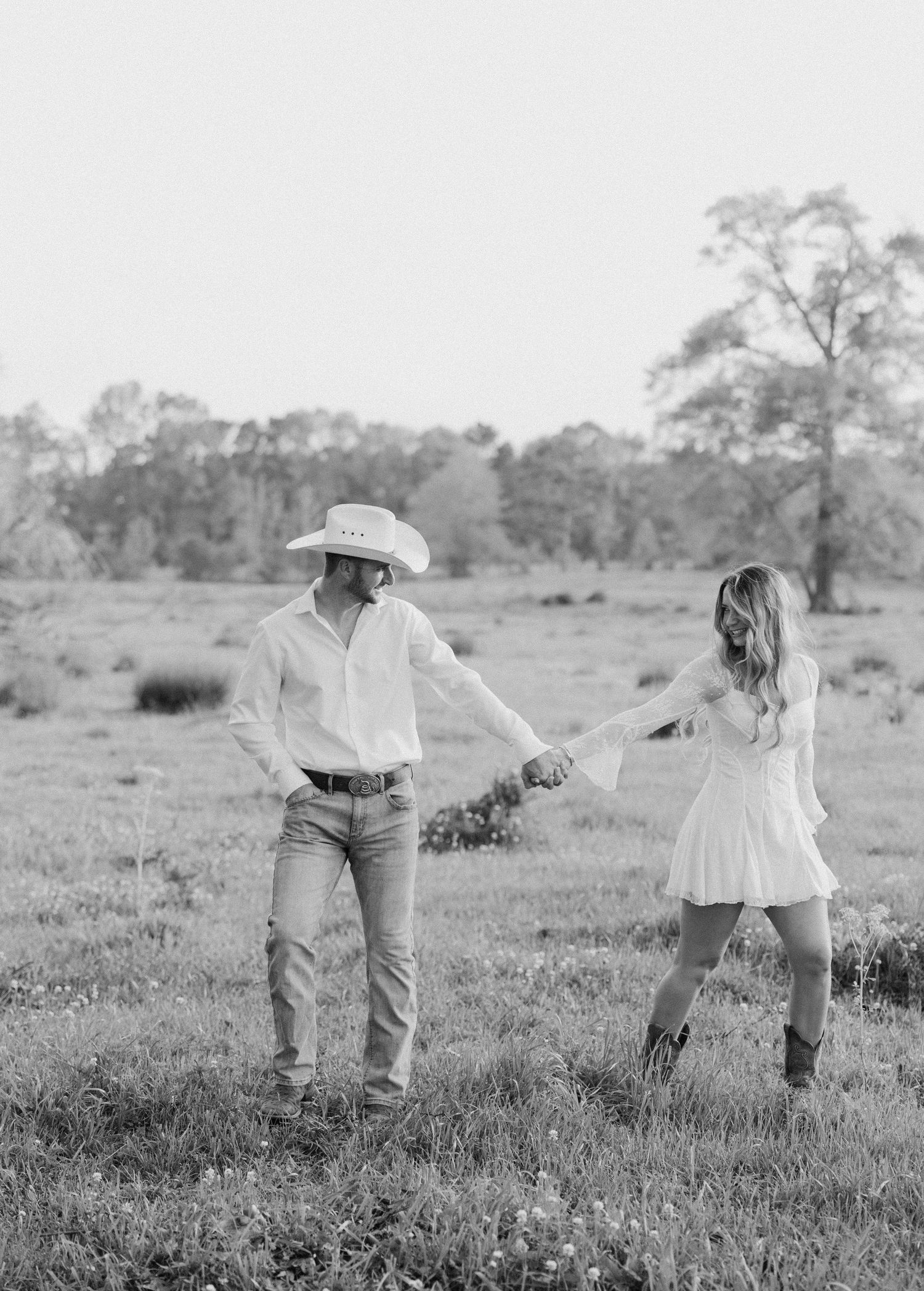 Black and white photo of a couple holding hands in a field, man wearing cowboy hat and woman in white dress with boots.