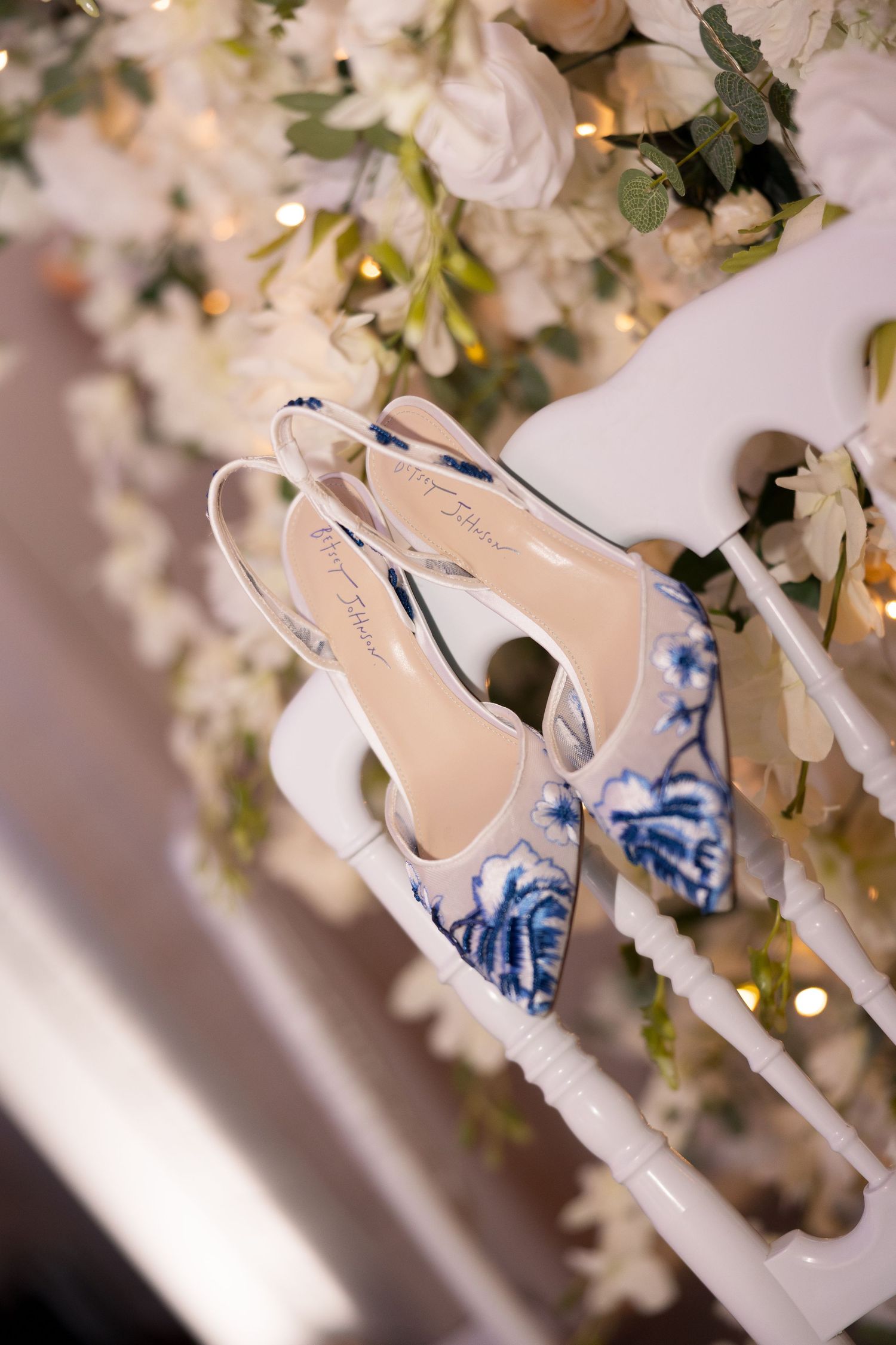 Blue and white floral embroidered bridal heels displayed on white chair rail with white roses and fairy lights backdrop.