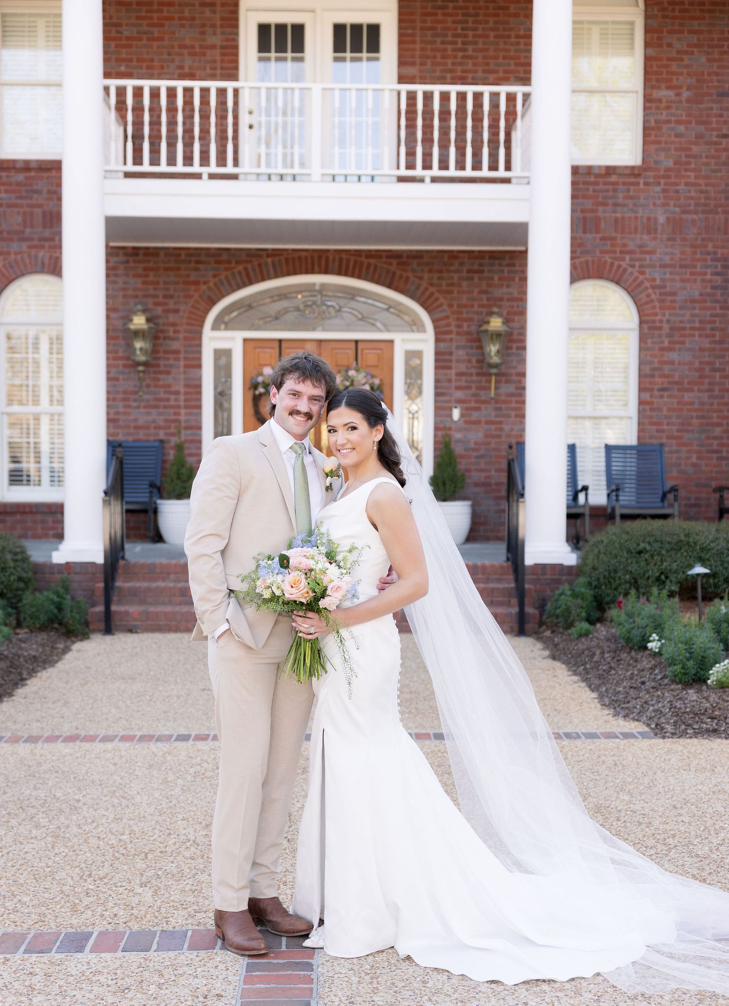 Bride and groom posing in front of elegant brick mansion, bride holding floral bouquet in white gown with veil.