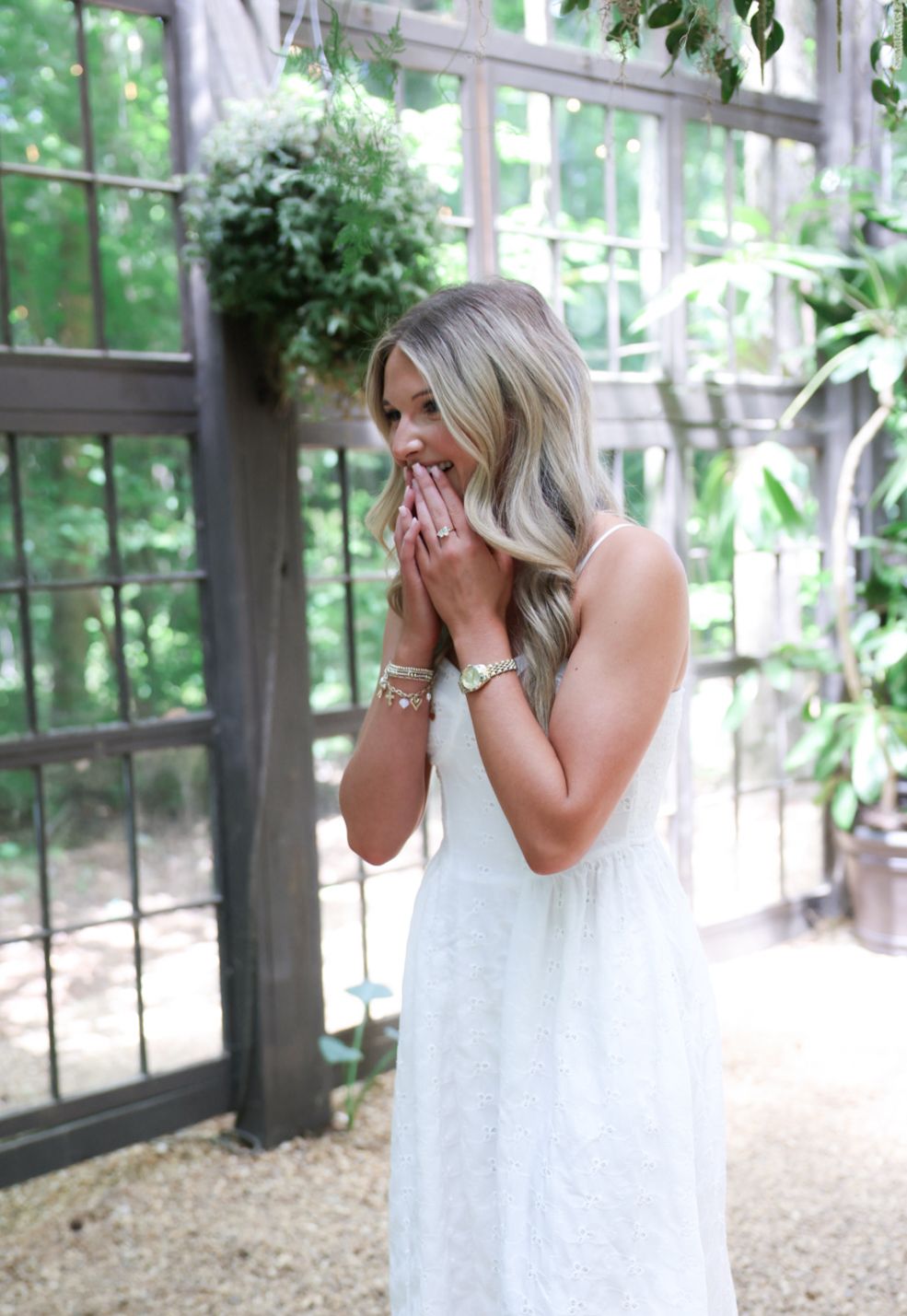 Woman in white lace dress covers mouth with hands in surprise, standing outdoors near rustic wooden structure.