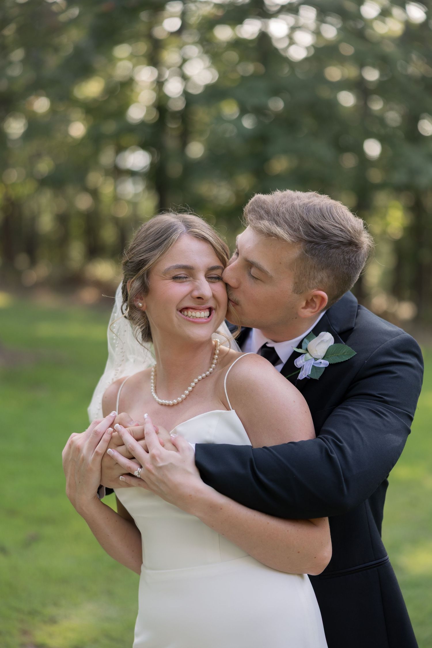 Groom in black suit kisses bride's cheek as she smiles, wearing white gown, pearl necklace, and veil outdoors.