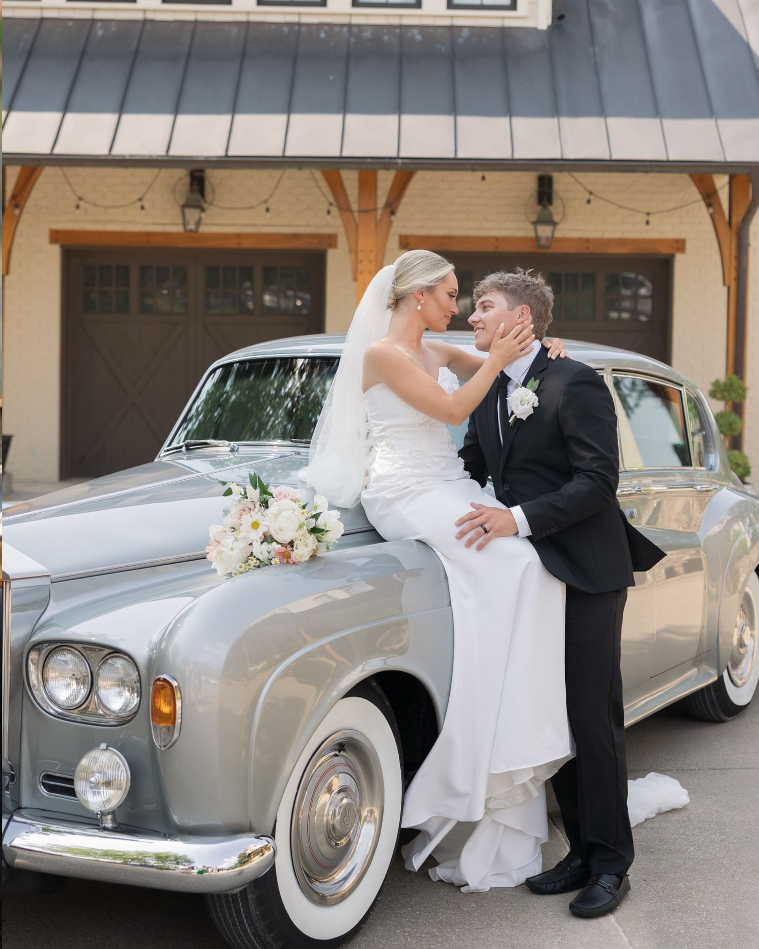 Bride and groom sharing a romantic kiss beside a classic silver Rolls-Royce on their wedding day.