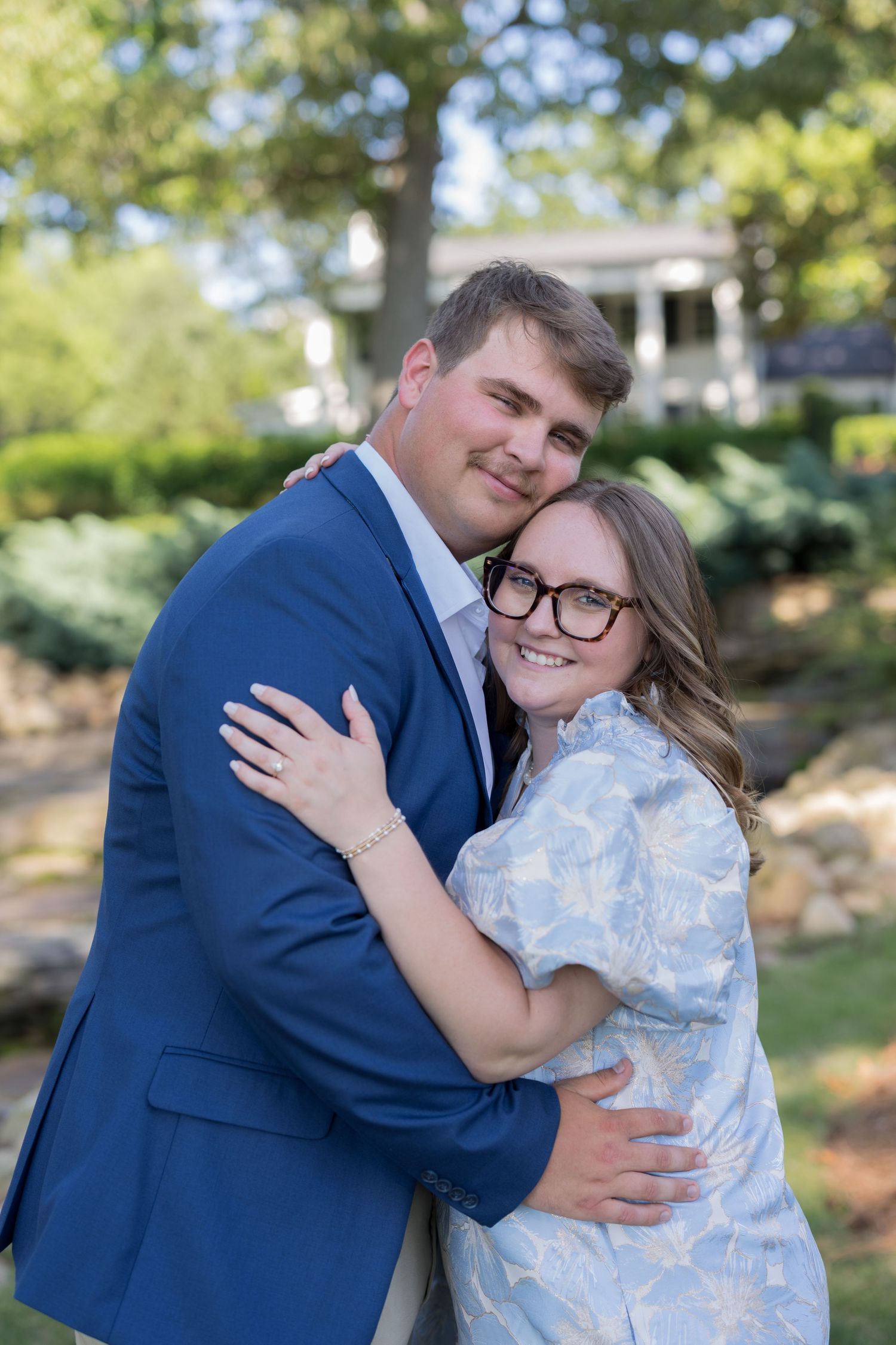 Couple embracing outdoors, man in blue suit and woman in floral dress with glasses, smiling in a garden setting.