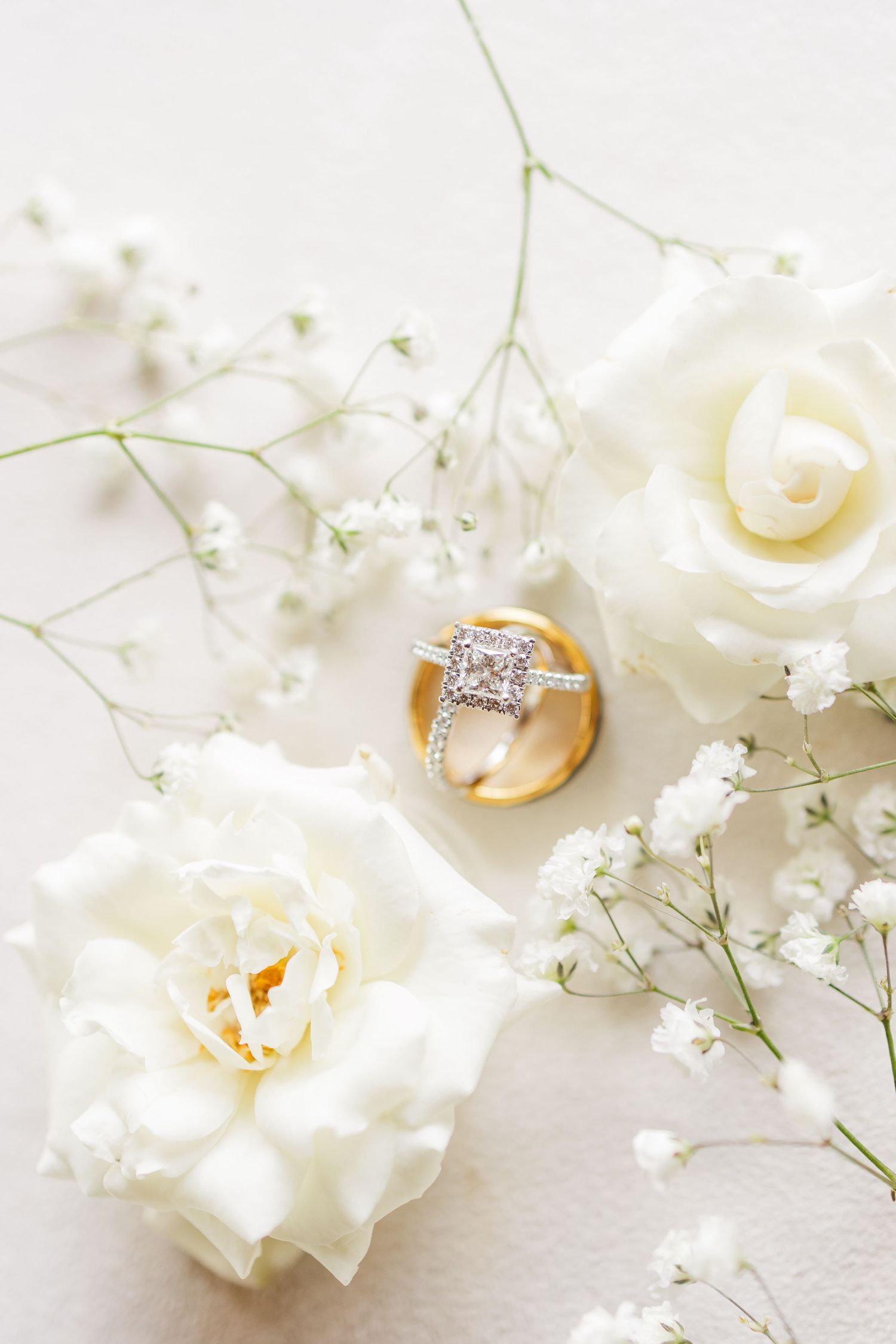 Diamond engagement ring and gold wedding band nestled among white roses and baby's breath on a soft white background.