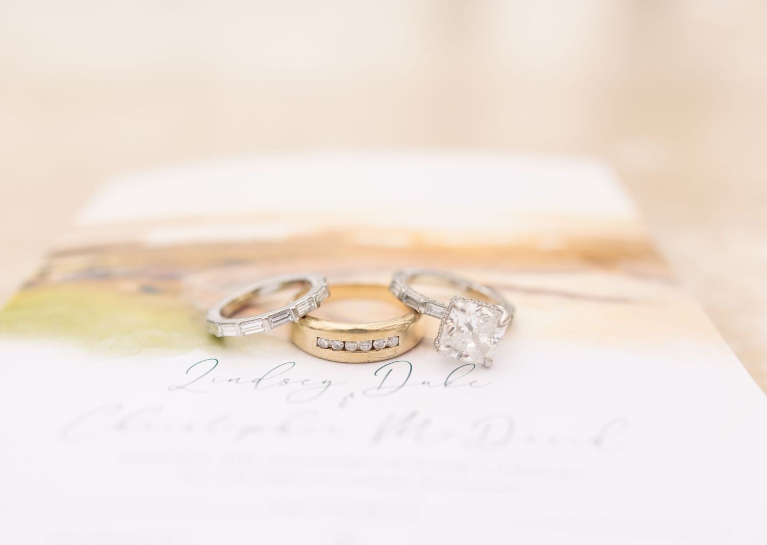 Three wedding rings, two silver and one gold, displayed on a wedding invitation with soft bokeh background.