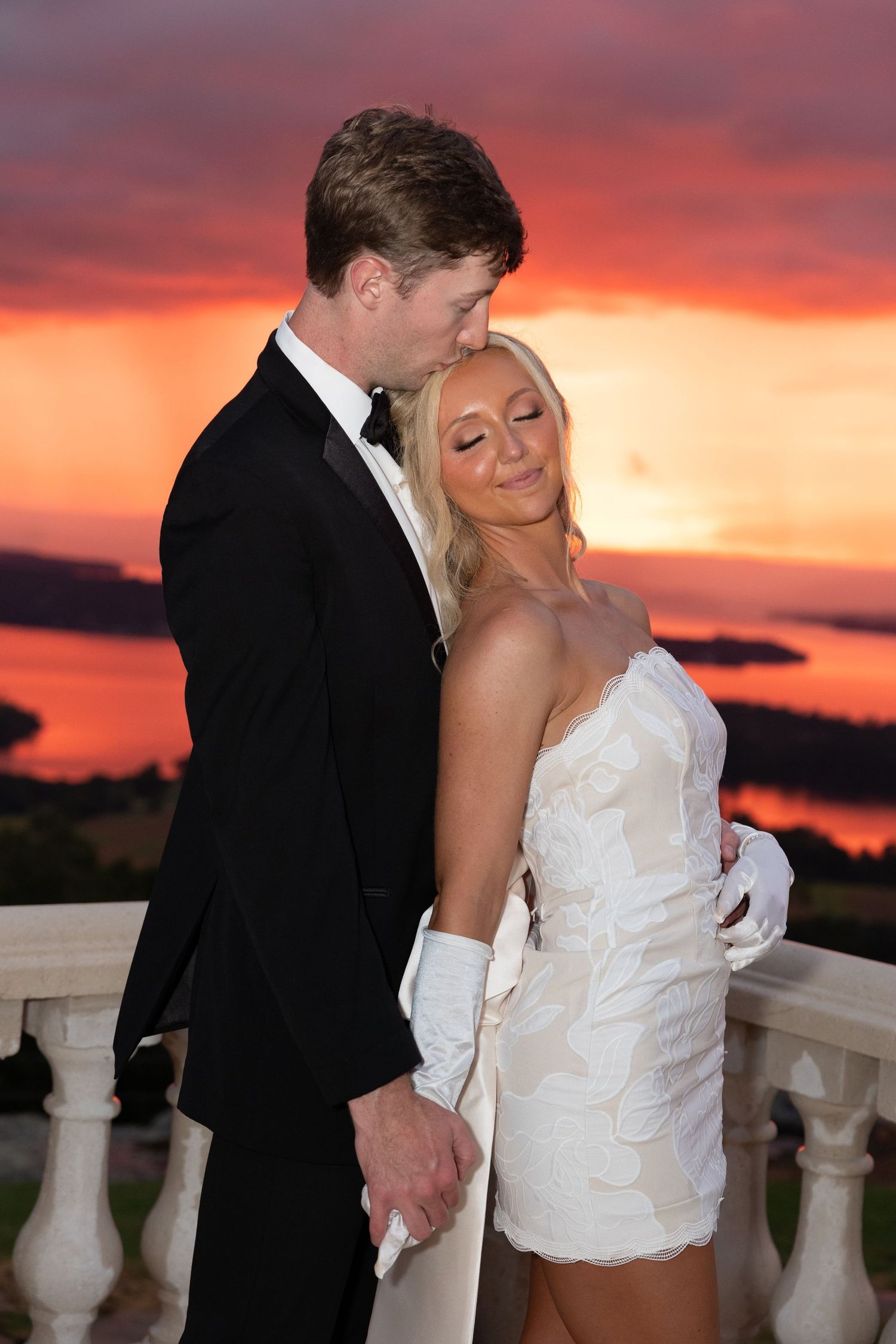 Bride and groom embracing on a balcony during a stunning red and orange sunset over a lake.