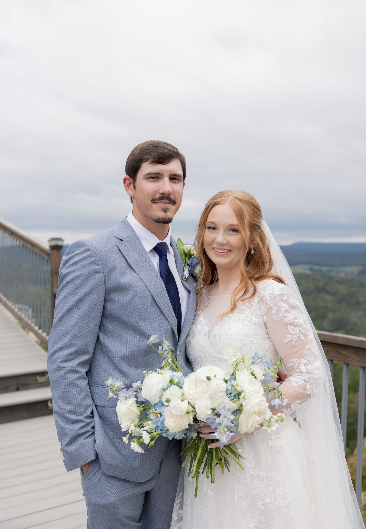 Bride and groom posing on outdoor deck with scenic view, bride holding white and blue bouquet, groom in gray suit.