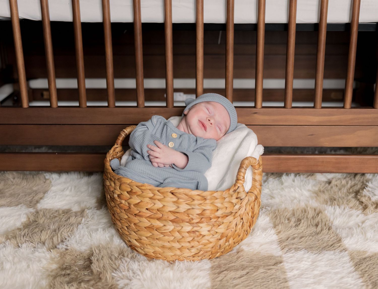 Newborn baby sleeping peacefully in a woven basket wearing a gray outfit and hat, placed in front of a wooden crib.