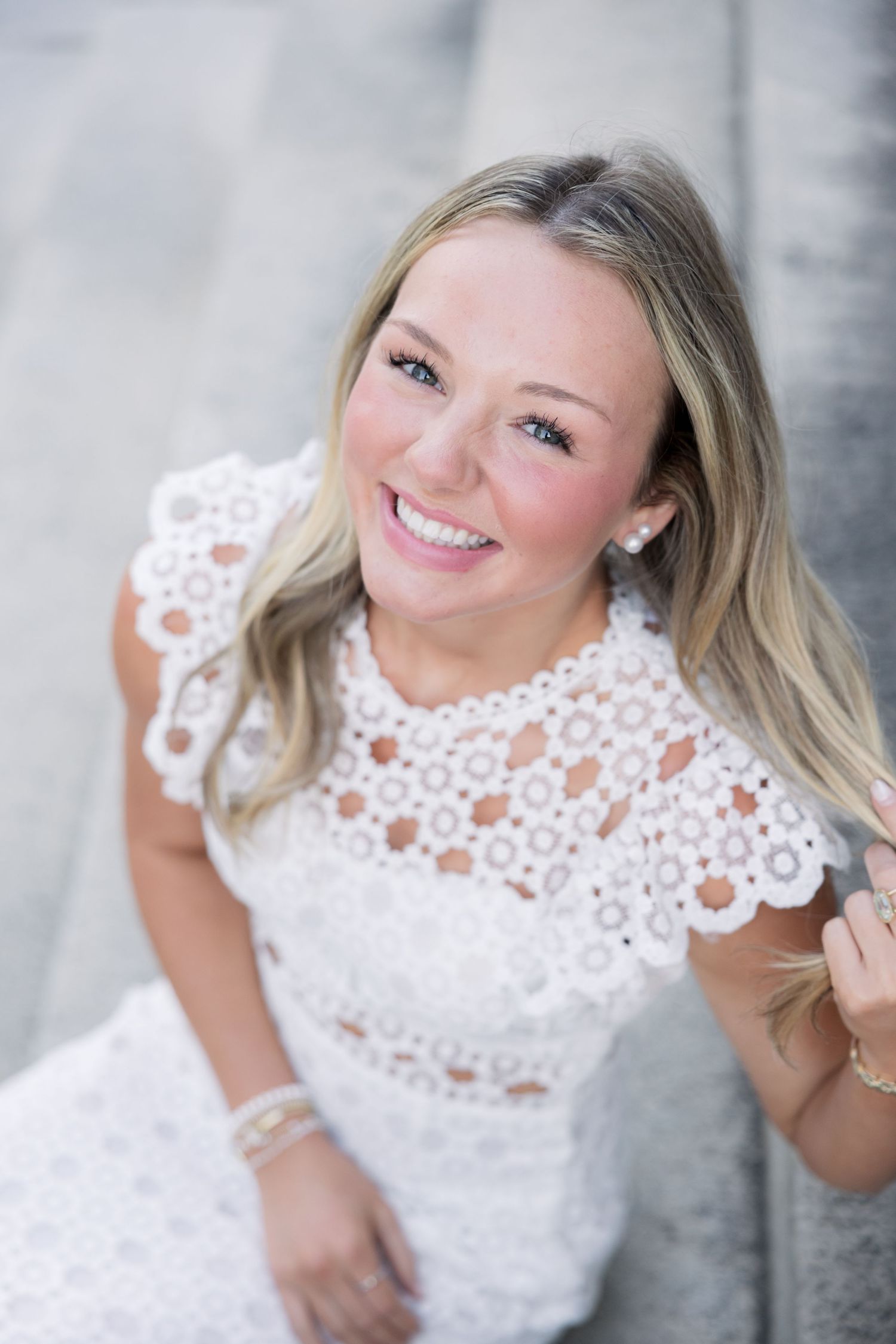 Smiling blonde woman in white lace dress looking up at camera outdoors, wearing pearl earrings and bracelet.