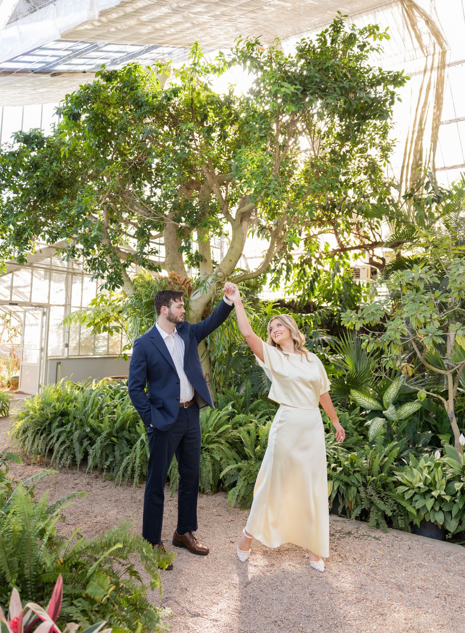 Couple dancing in a lush greenhouse, man in navy suit twirling woman in ivory dress among tropical plants.