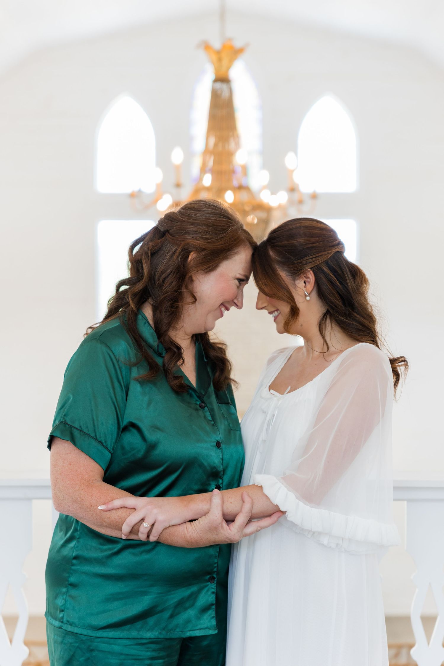 Bride in white gown and woman in green satin pajamas share a tender forehead-to-forehead moment in elegant white chapel.