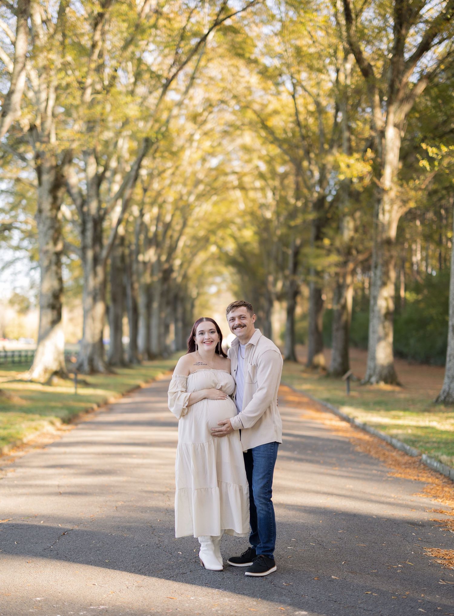 Couple poses on tree-lined path during golden hour maternity photo shoot, woman in cream dress cradling baby bump.