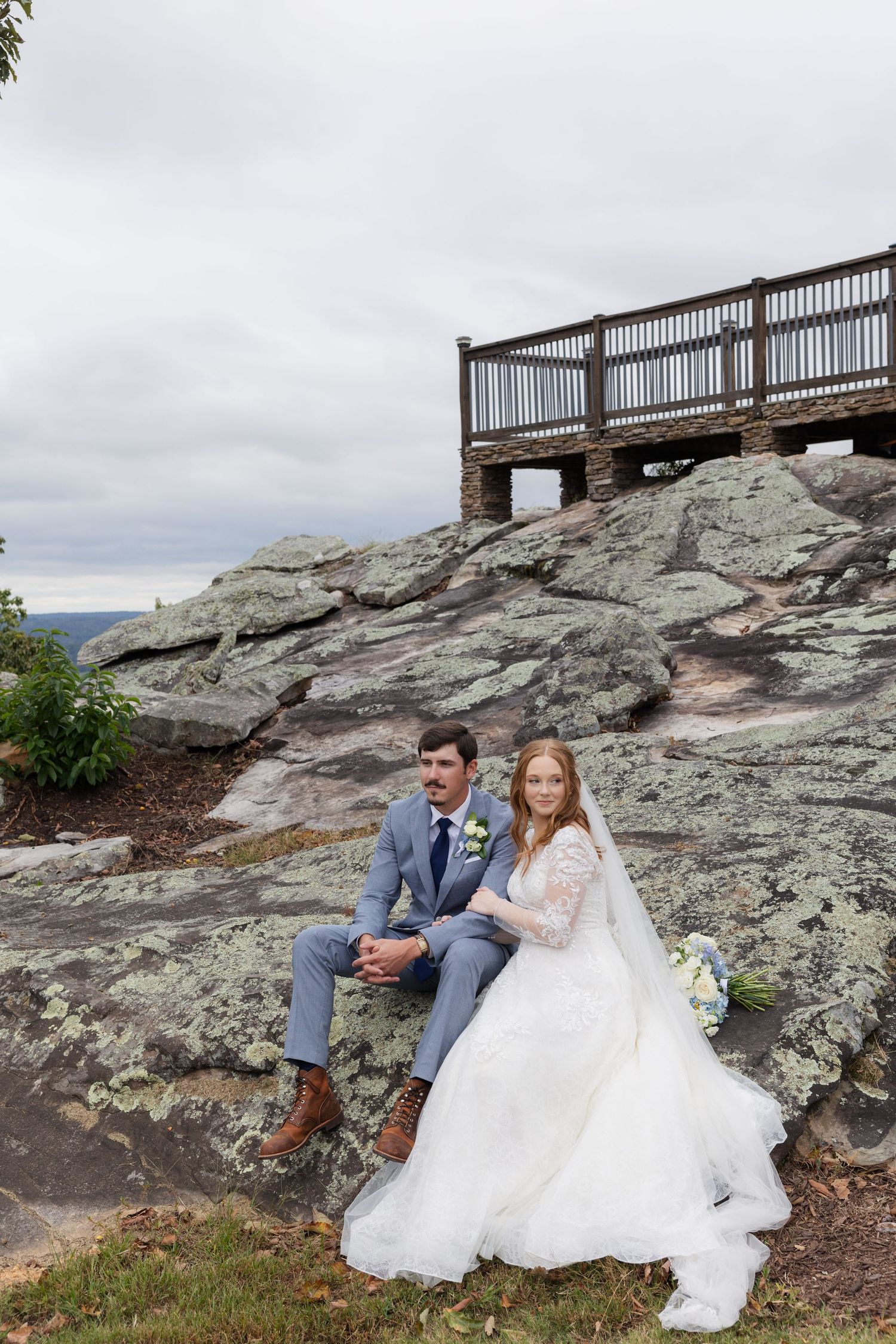 Bride and groom sitting on rocky outcrop in wedding attire, wooden overlook deck behind them under cloudy sky.