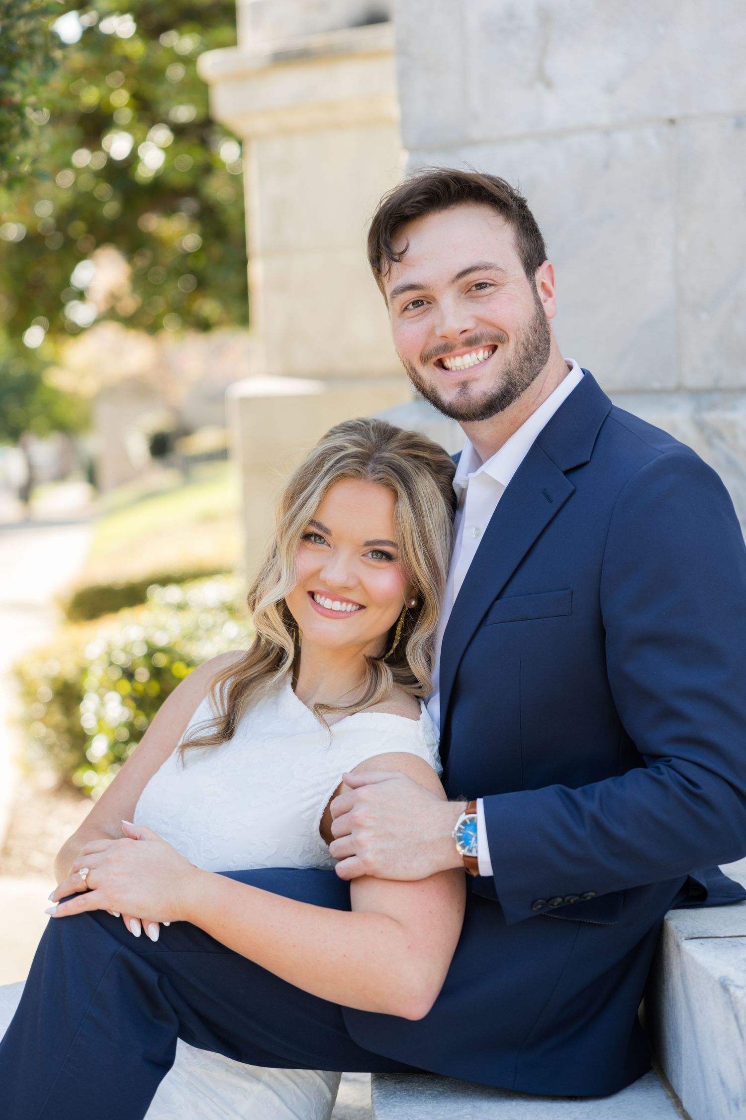 A smiling couple poses together outdoors, the woman in white and man in a navy suit, sitting by stone columns.