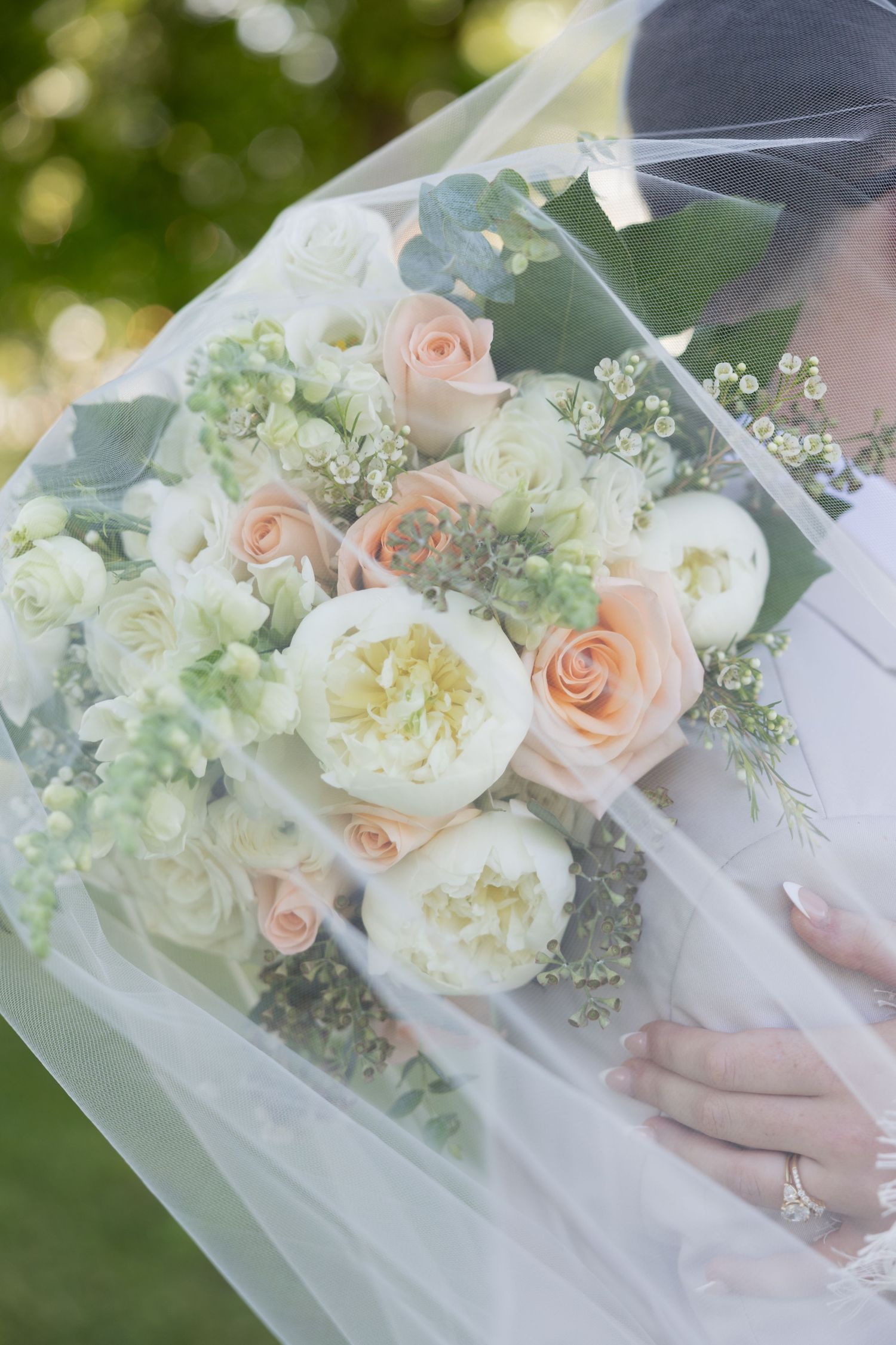 Bride holding a lush wedding bouquet of white peonies and peach roses under a sheer veil outdoors.