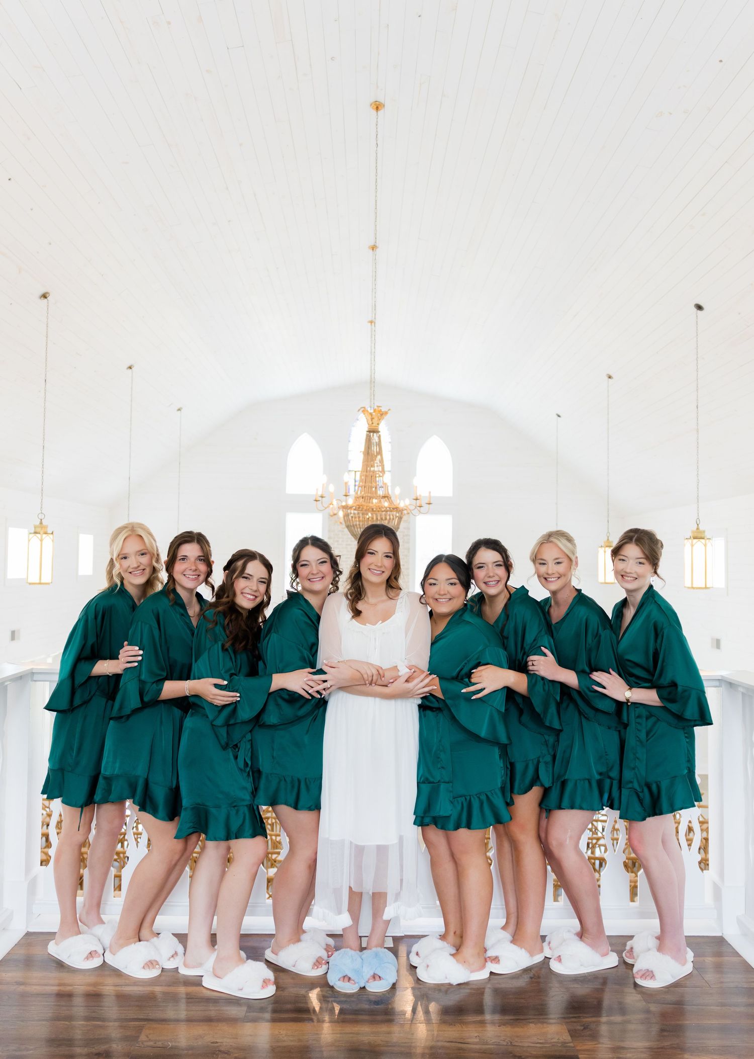 Bride in white gown poses with eight bridesmaids in emerald green robes and white slippers inside elegant white chapel.
