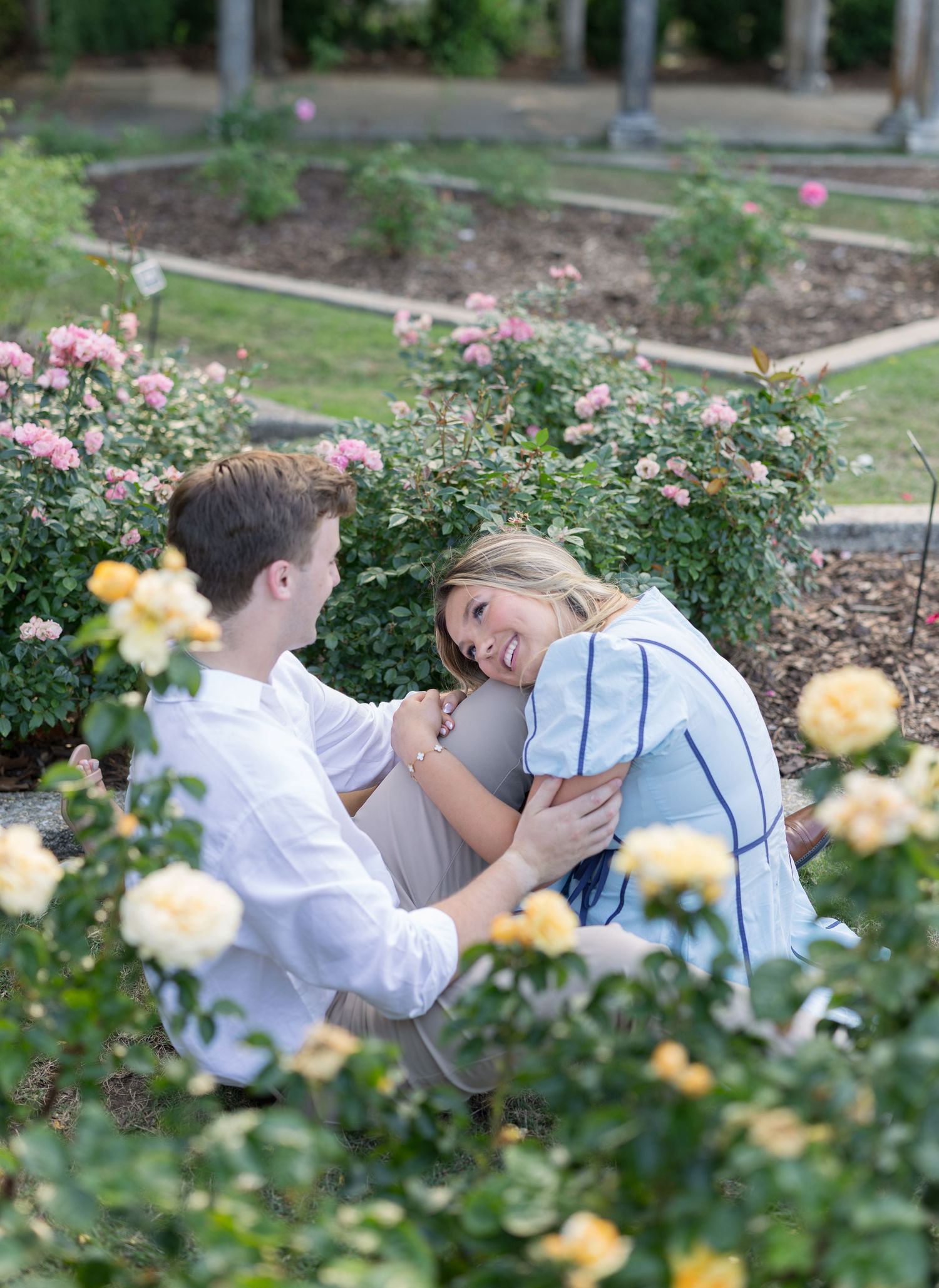 Couple sitting together in a rose garden, smiling and embracing surrounded by pink and yellow blooming roses.