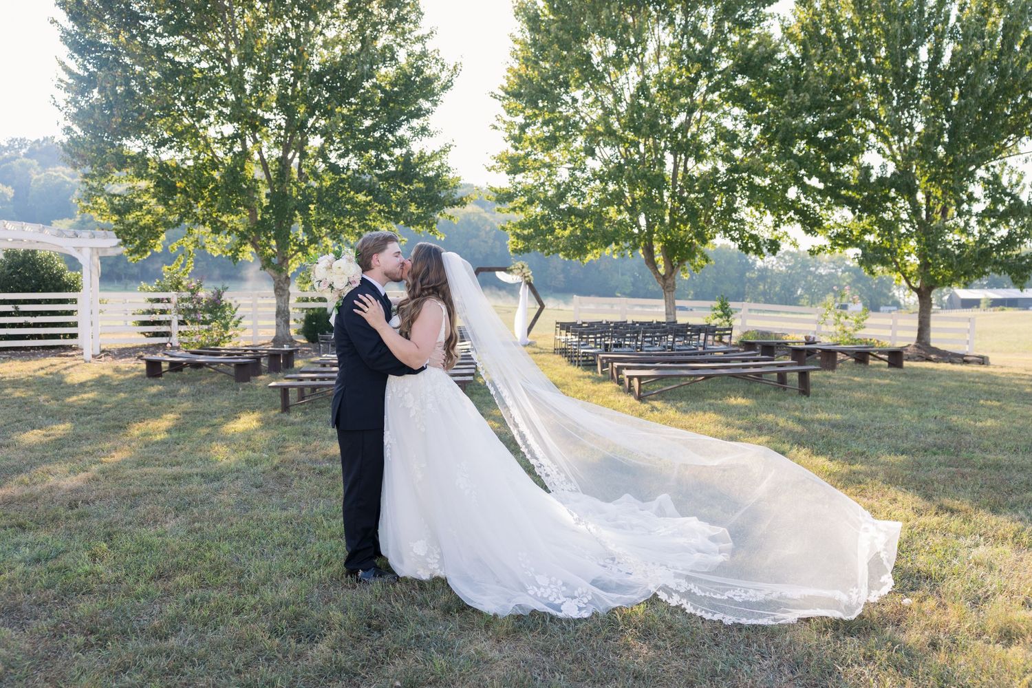 A bride and groom share a romantic kiss outdoors at a wedding venue, with the bride's veil flowing gracefully in the breeze.