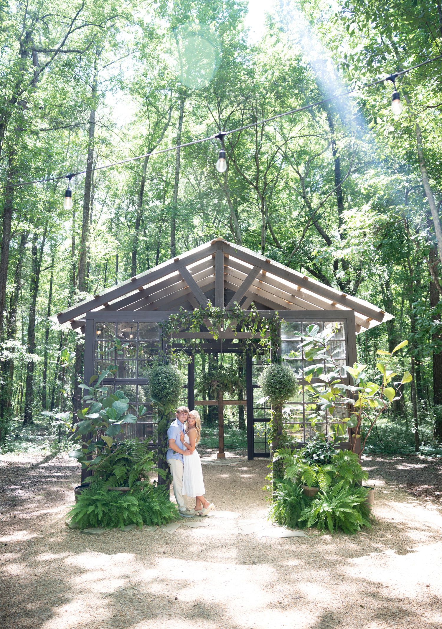 Couple embraces under a rustic wooden gazebo adorned with greenery and ferns in a sunlit forest setting.