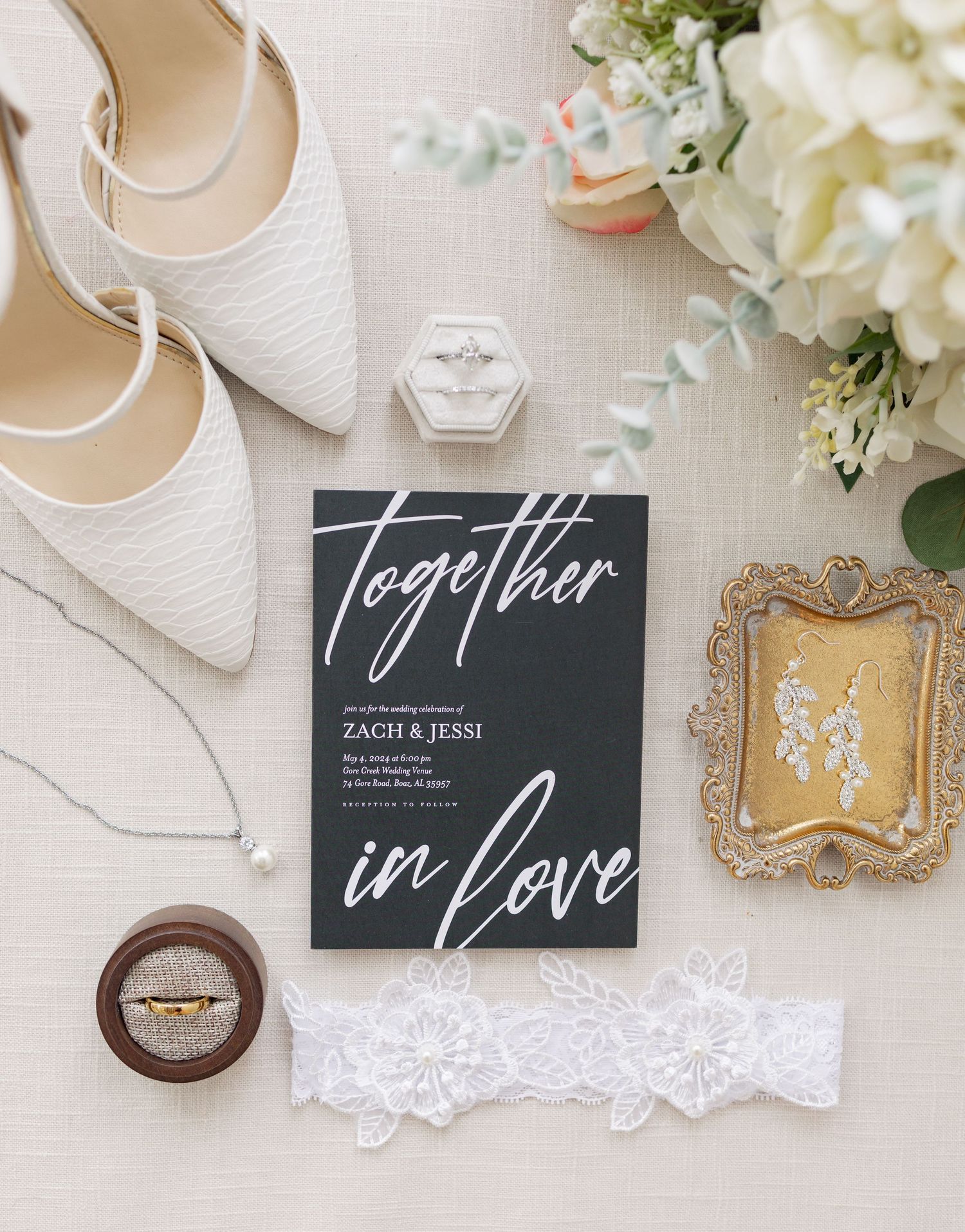 Elegant wedding flat lay featuring white heels, floral bouquet, ring box, lace garter, and a black Together in Love invitation.