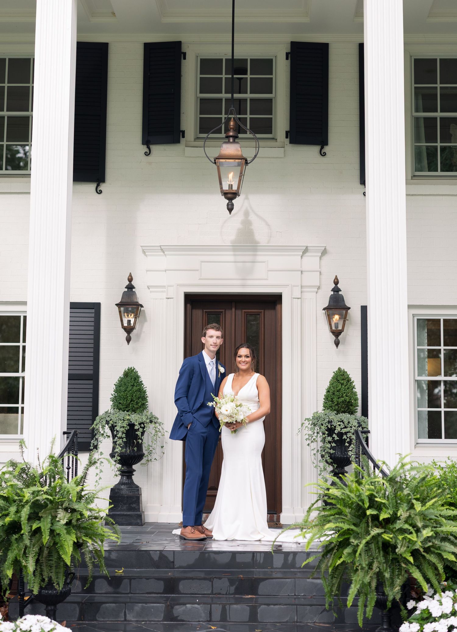 Bride and groom pose on steps of elegant white colonial mansion with black shutters and lush greenery on their wedding day.