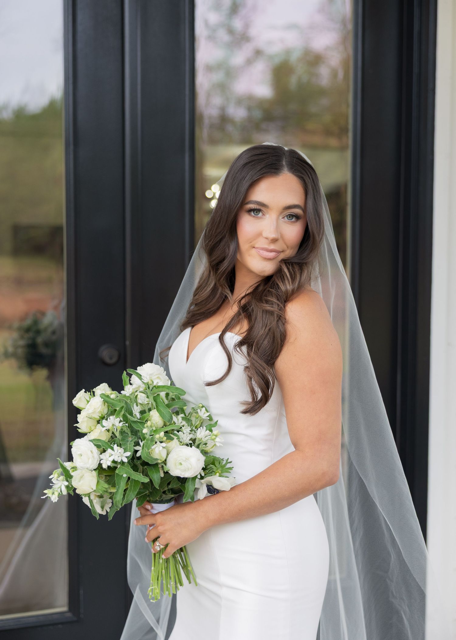Bride in white gown holding white floral bouquet, wearing veil, posing elegantly near glass doors.