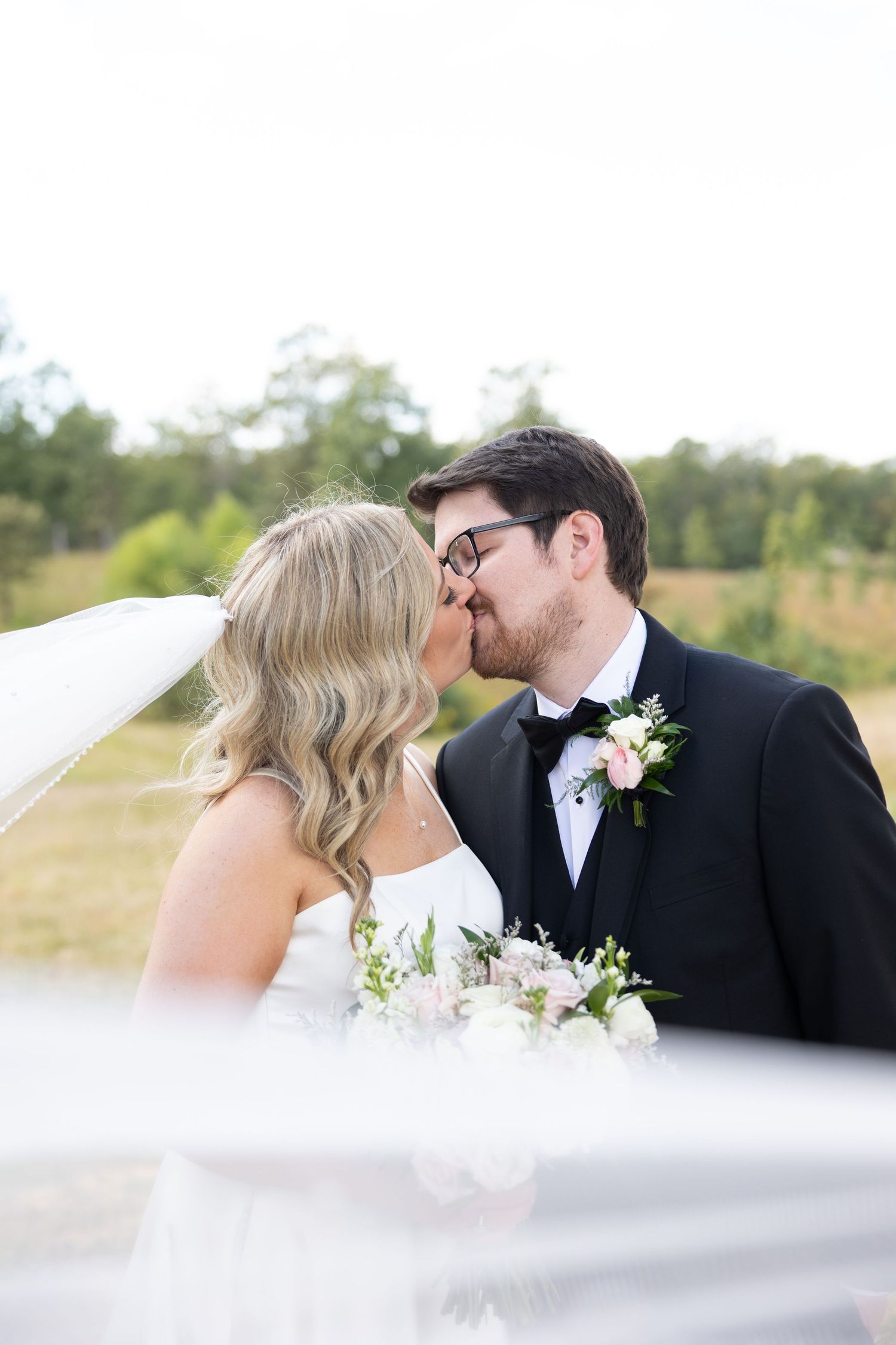 Bride and groom share a kiss outdoors, bride holding pink bouquet, groom in black tuxedo with floral boutonniere.