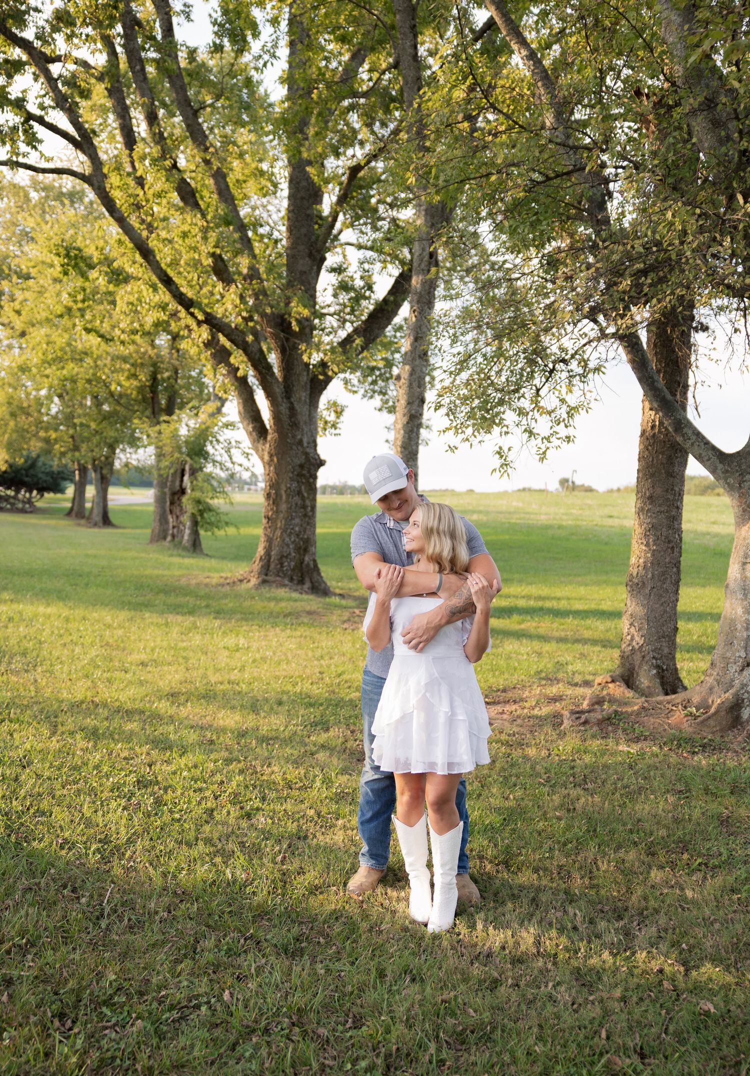 Couple embracing and kissing outdoors under green trees on a sunny day, woman in white dress and boots.