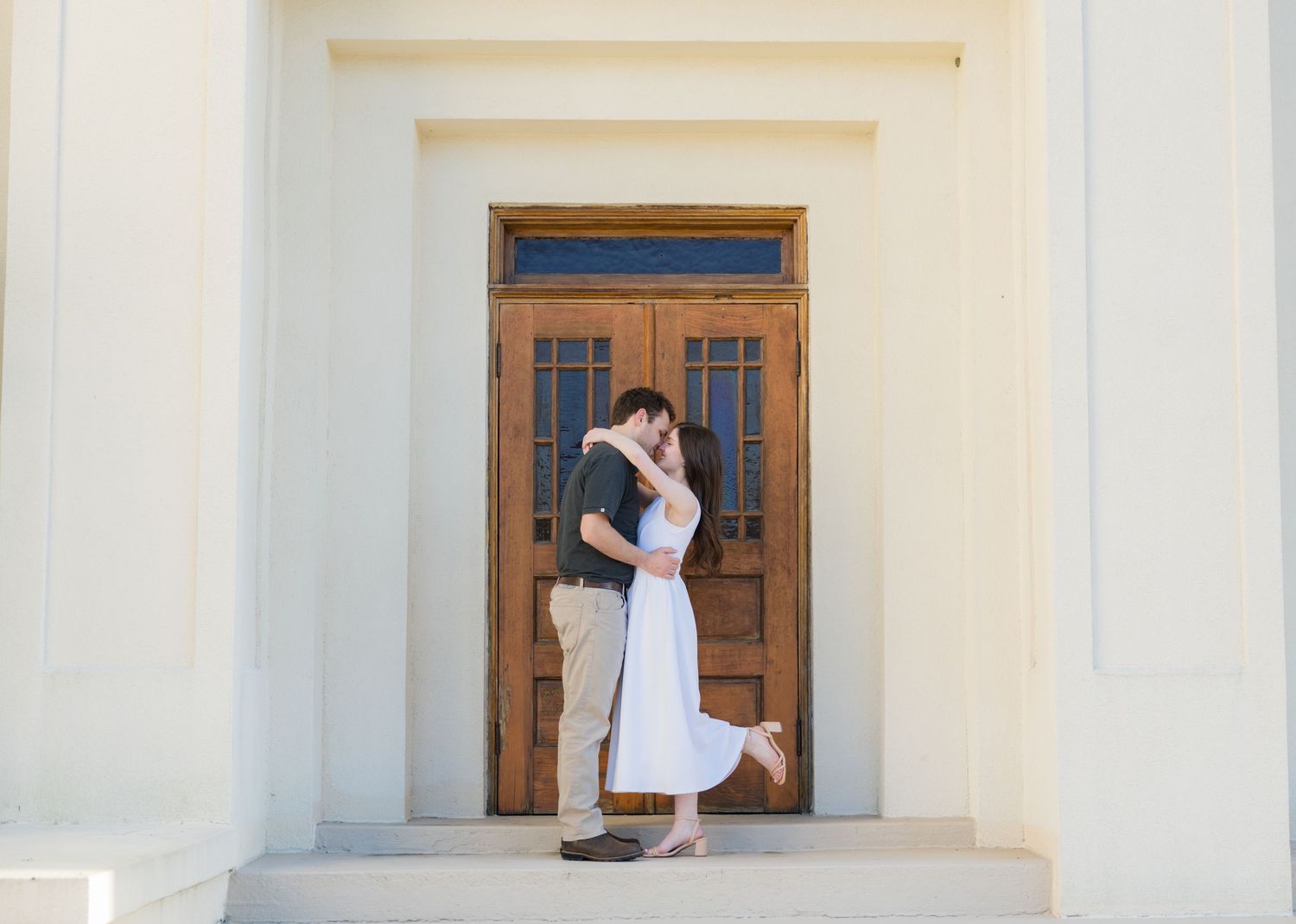 Couple sharing a romantic kiss in front of elegant wooden doors at a white classical building.