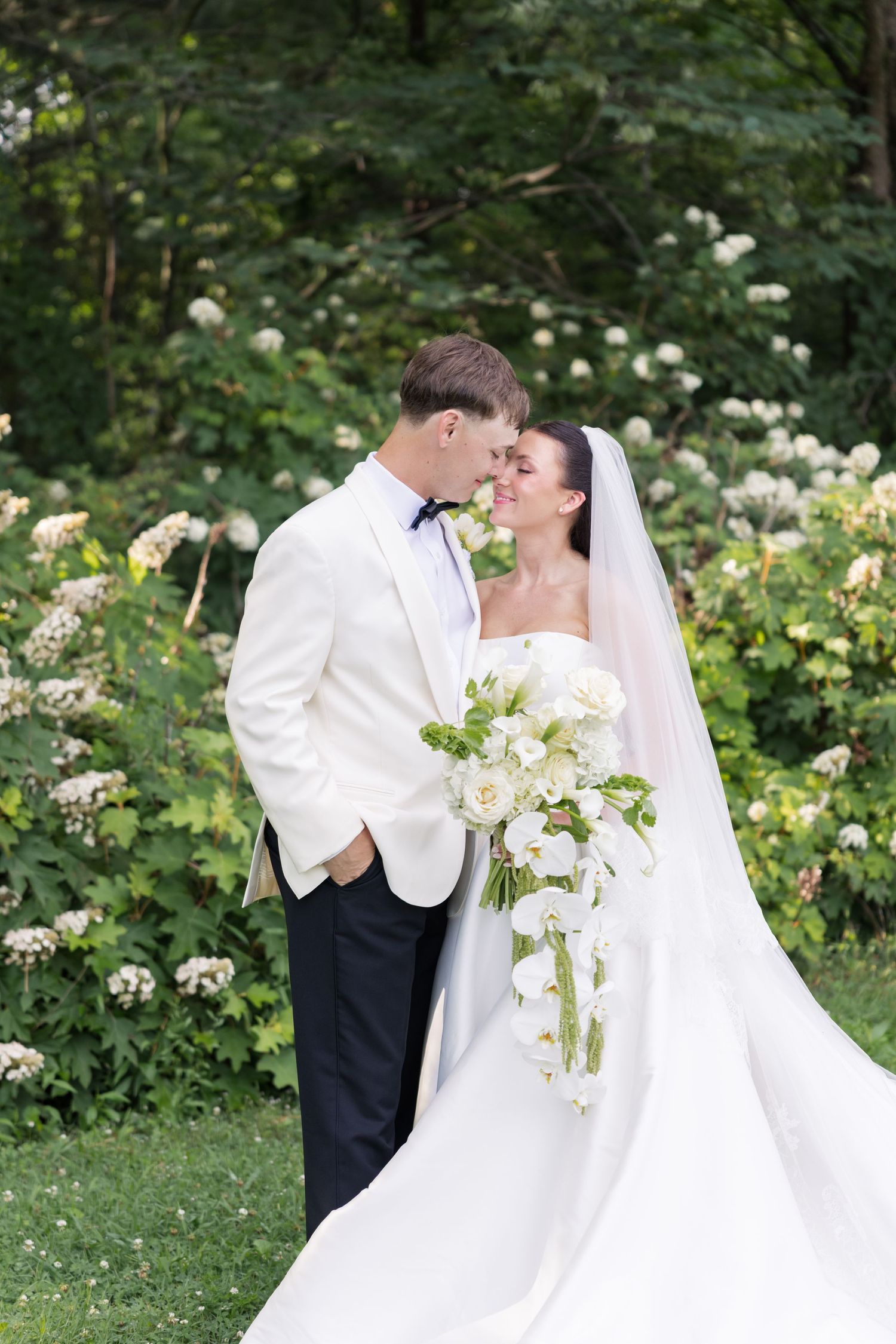 Bride and groom share romantic moment outdoors, she holds white cascading bouquet, he wears white tuxedo jacket.