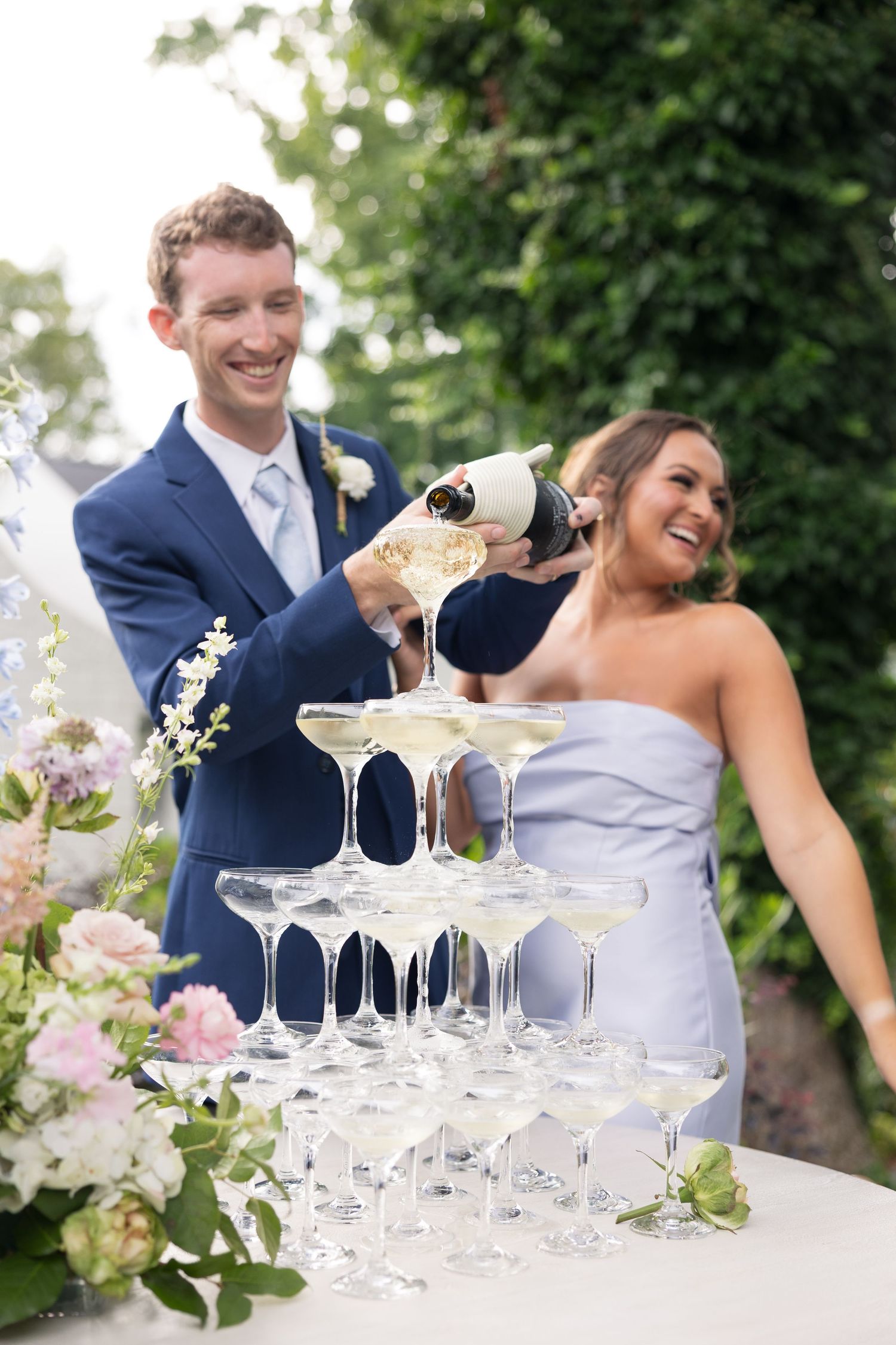 Smiling groom in blue suit pours champagne tower at outdoor wedding reception with laughing bridesmaid beside floral arrangements.