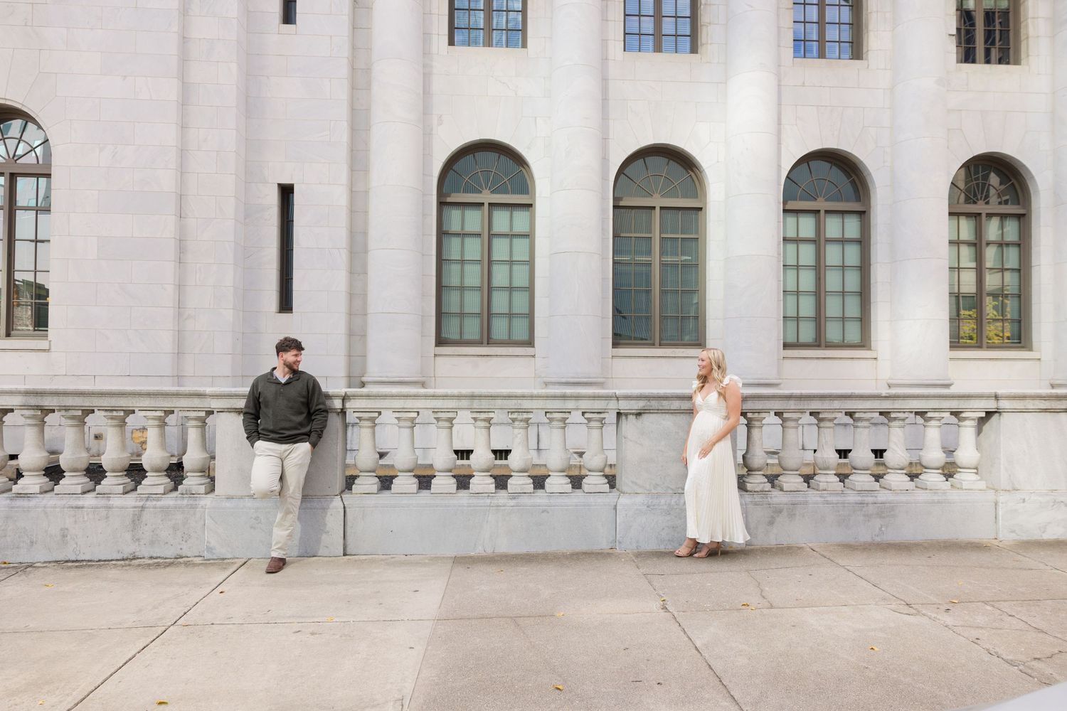 Couple posing apart by ornate stone building with arched windows for an engagement photo session.