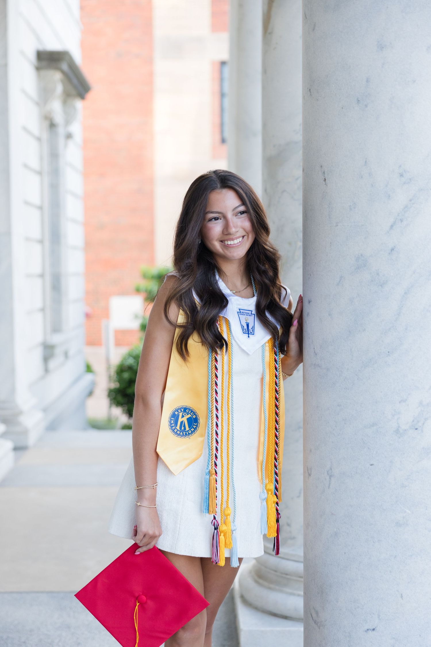 Smiling female graduate in white dress with gold honor stole and red cap poses by marble column at graduation ceremony.