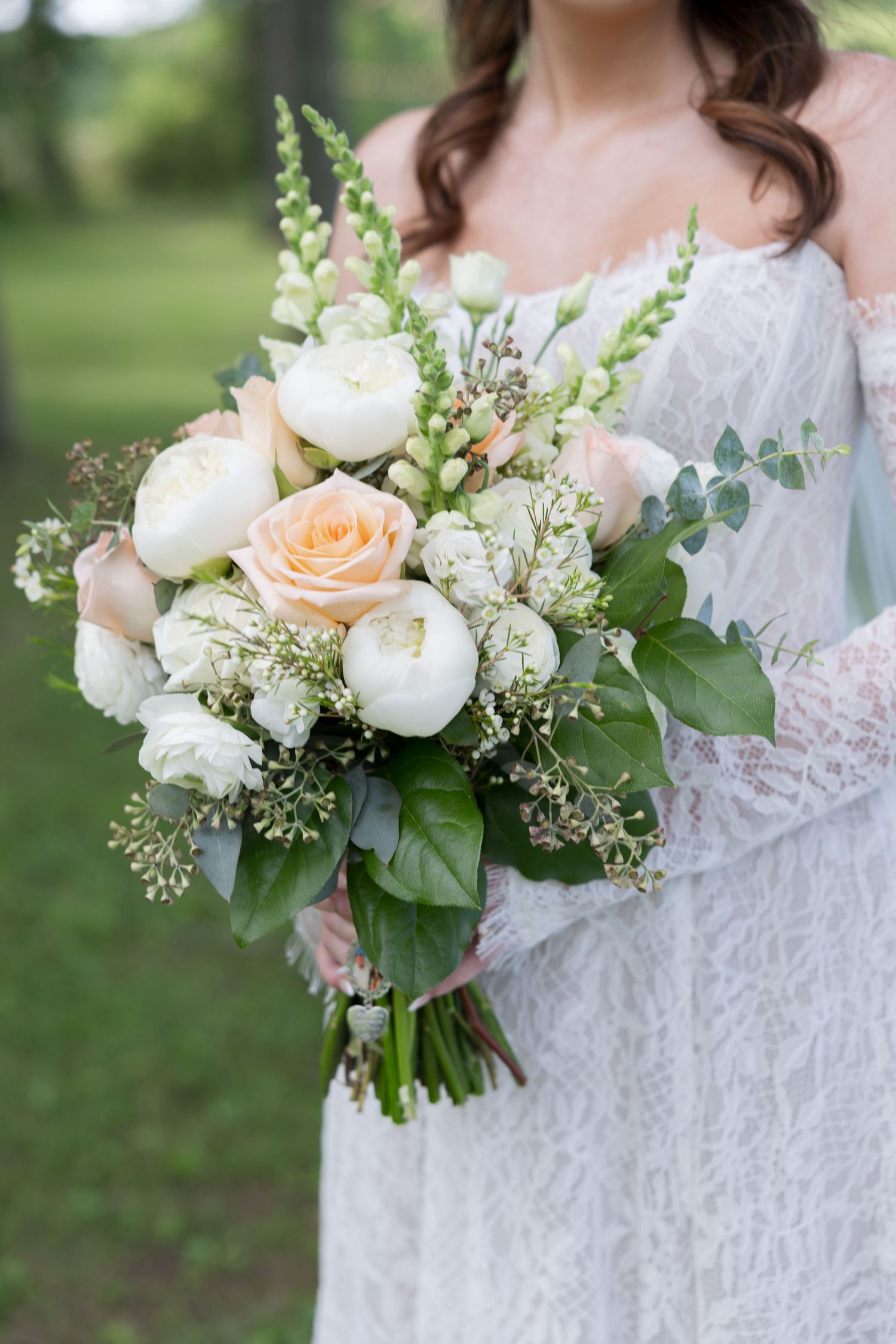 Bride in lace dress holding elegant wedding bouquet with white peonies, peach roses, and greenery.
