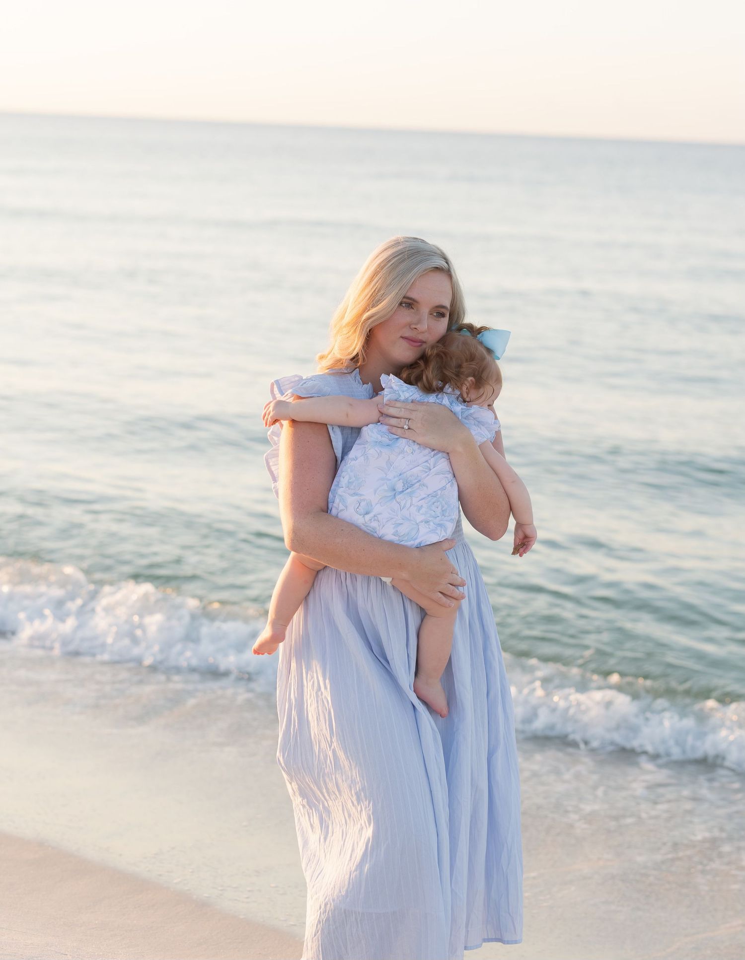 Blonde mother in light blue dress tenderly holds her baby girl on a serene sandy beach at sunset.