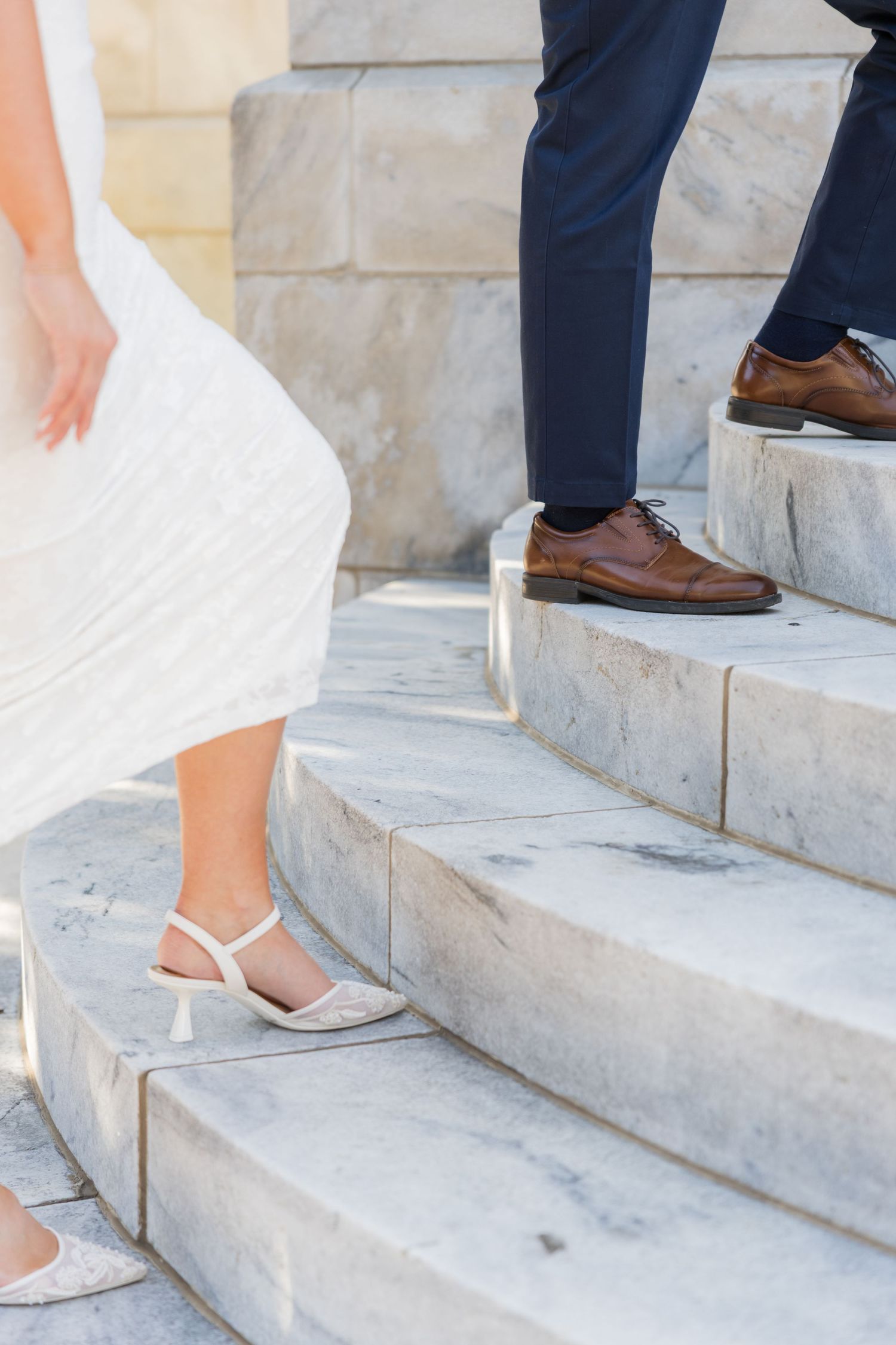 Bride in white dress and heels with groom in navy suit and brown leather shoes walking up marble steps.