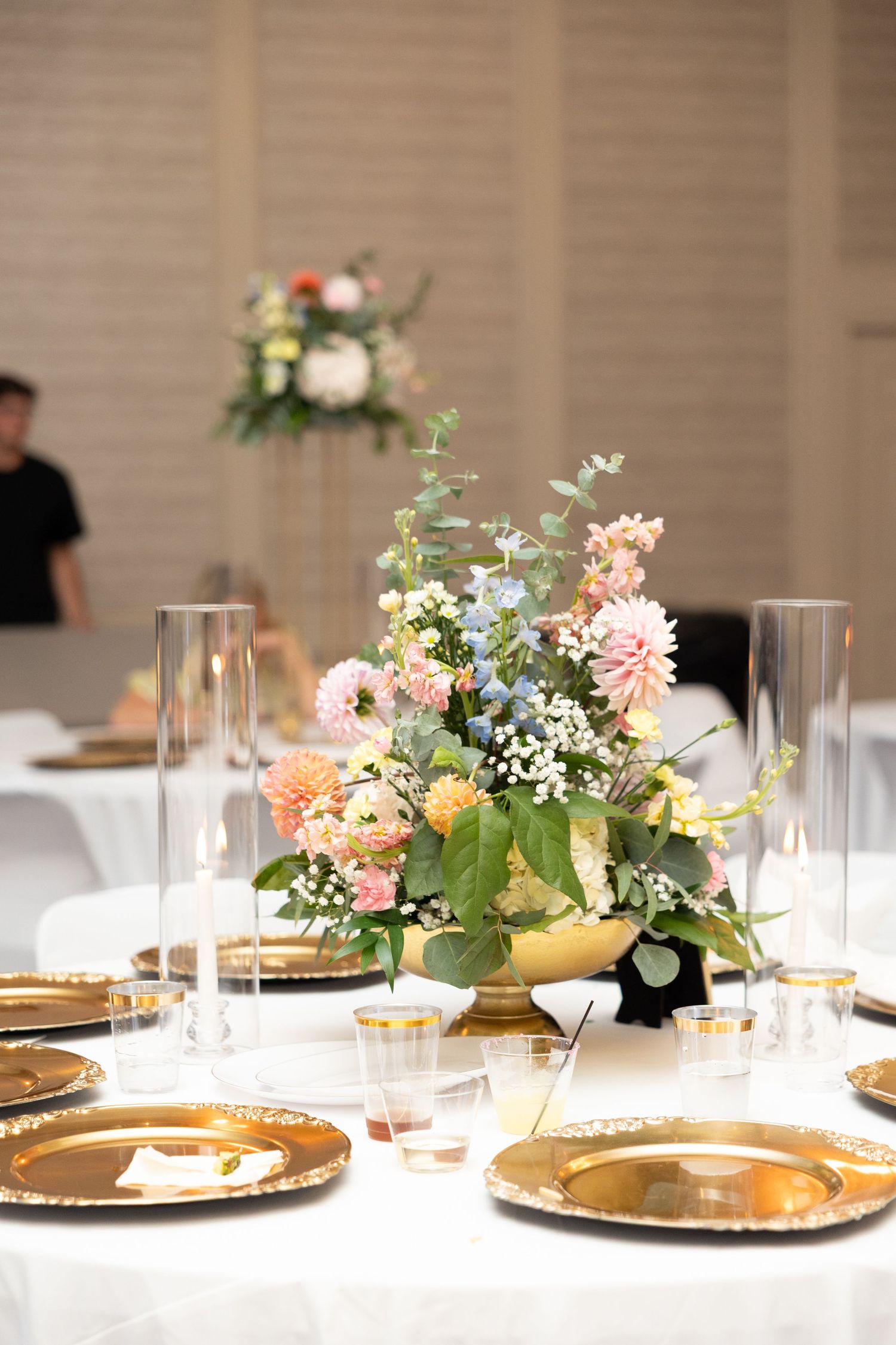 Elegant wedding reception table with gold charger plates, glass candleholders, and a pastel floral centerpiece in a gold bowl.