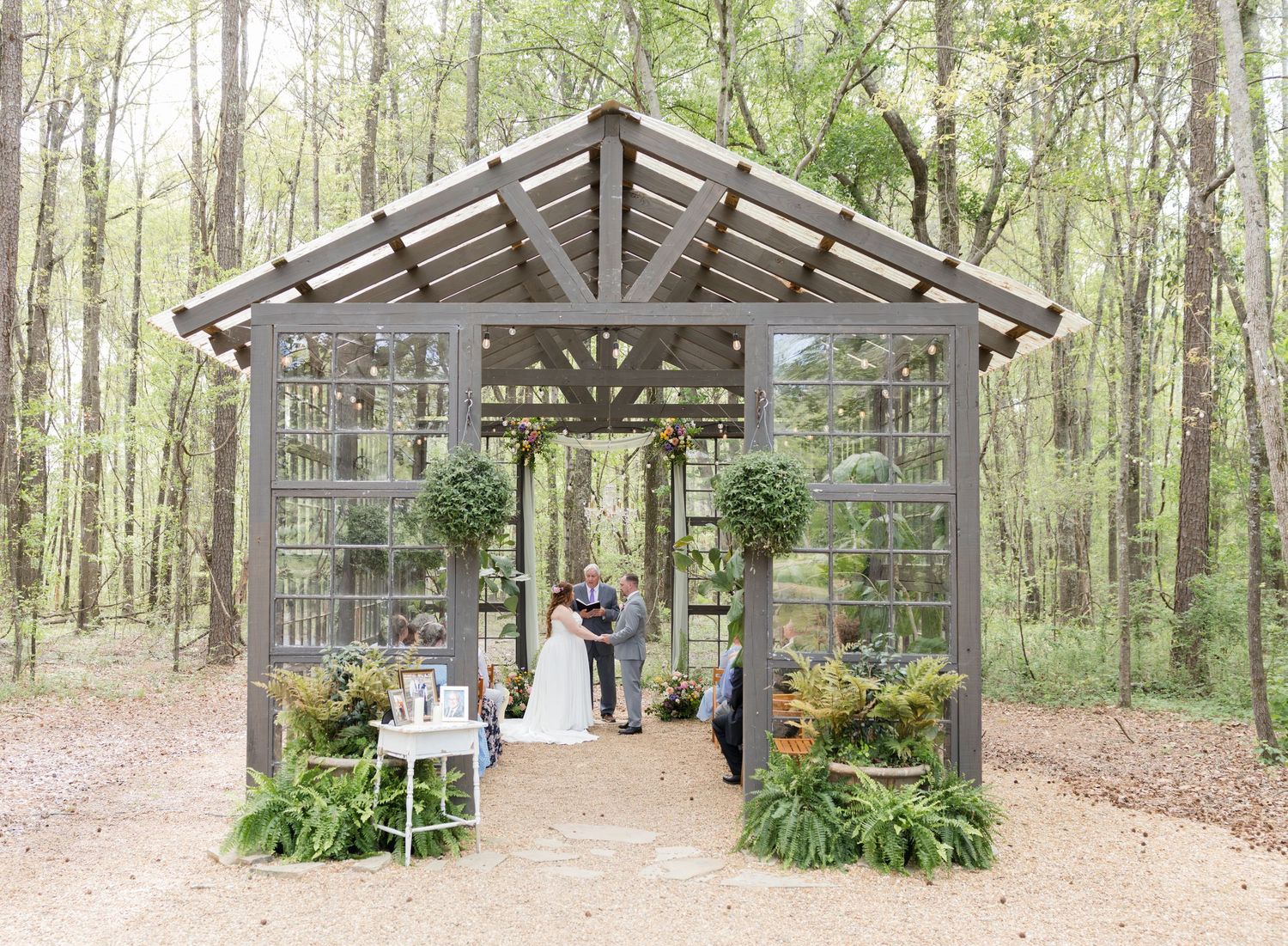 Rustic outdoor wedding ceremony in a glass greenhouse gazebo surrounded by lush green forest trees and fern decorations.