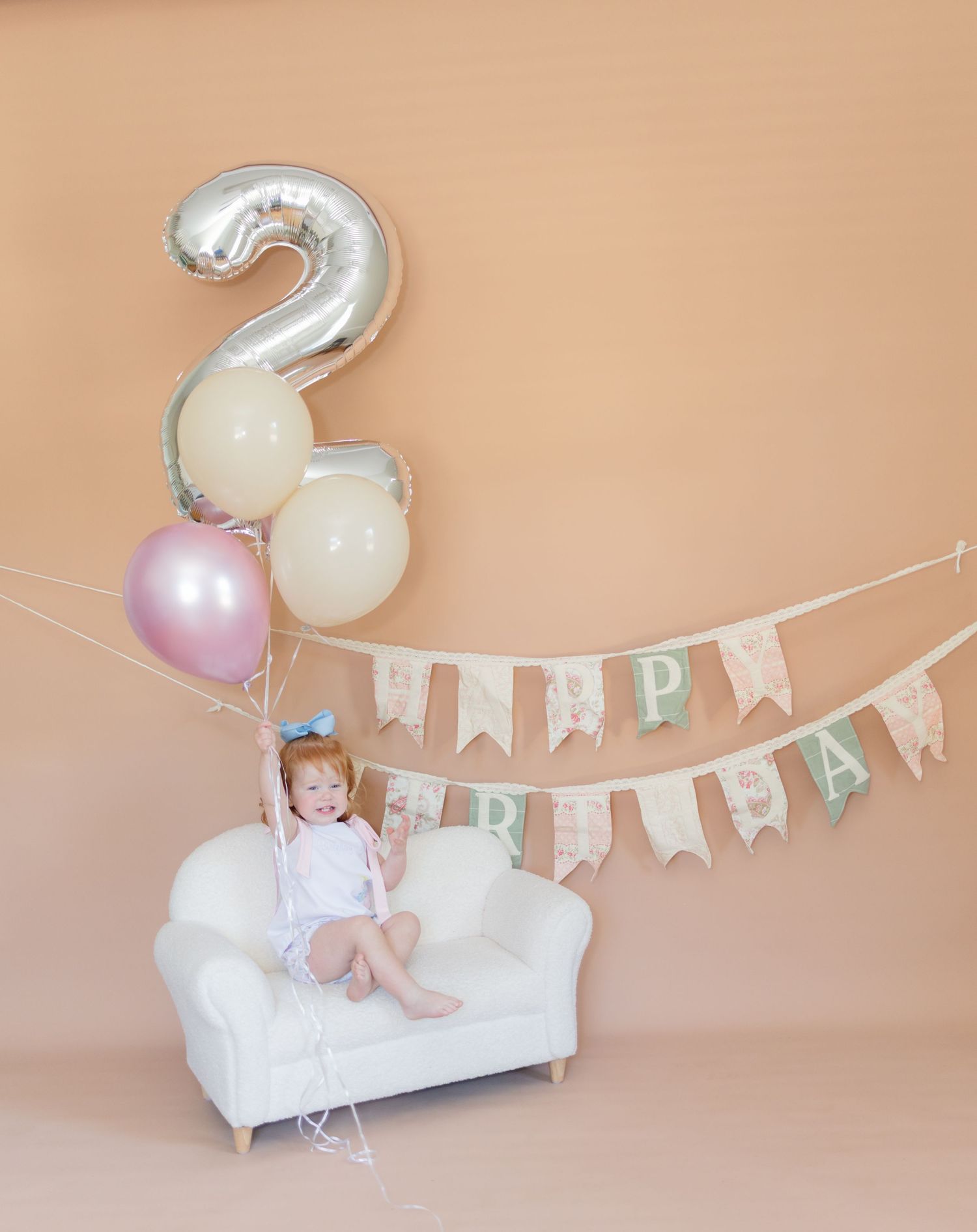 Toddler girl sitting on white mini sofa with birthday balloons and Happy Birthday banner celebrating her 2nd birthday.