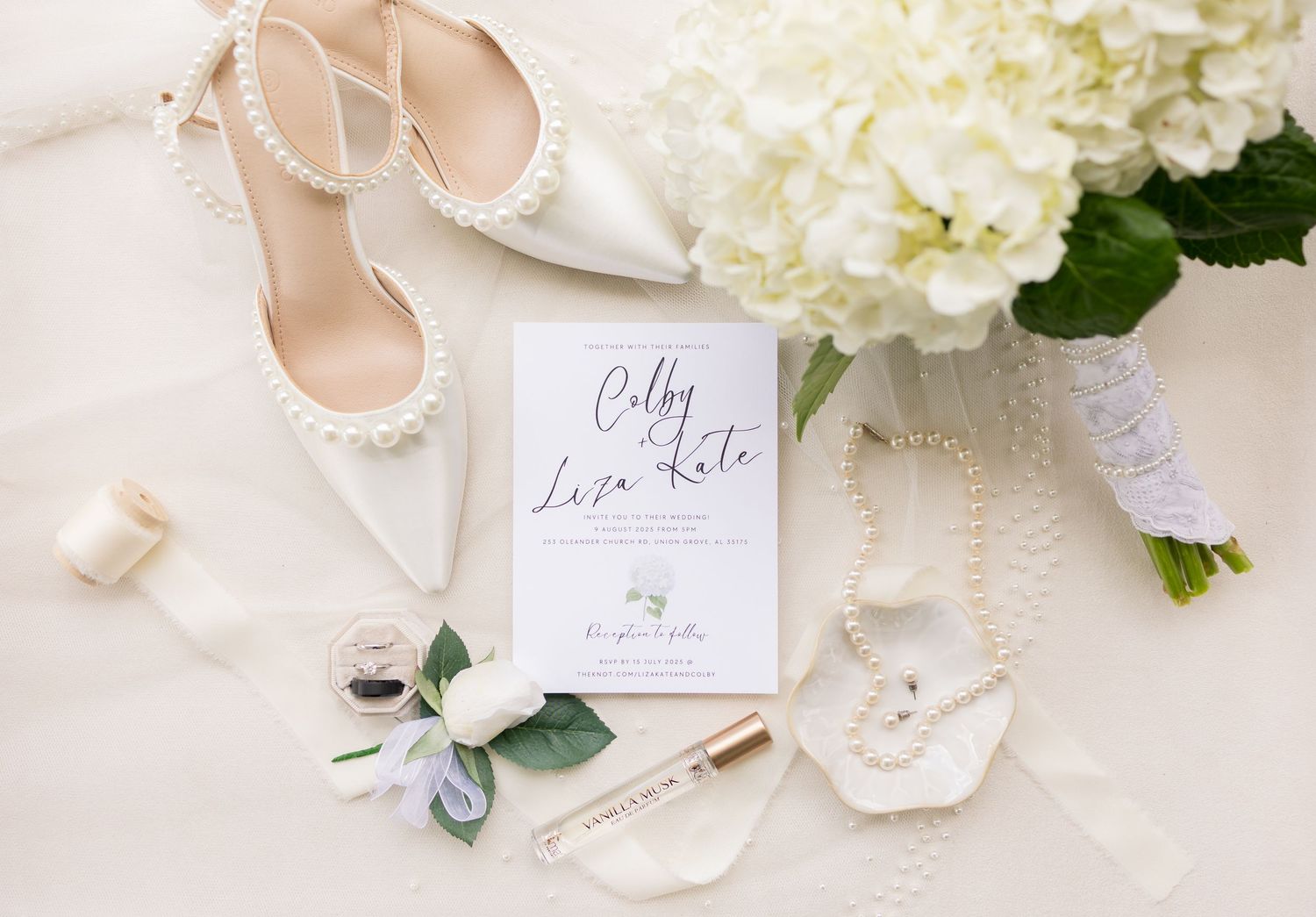 Elegant wedding flat lay with pearl heels, hydrangea bouquet, invitation, and jewelry on white fabric.