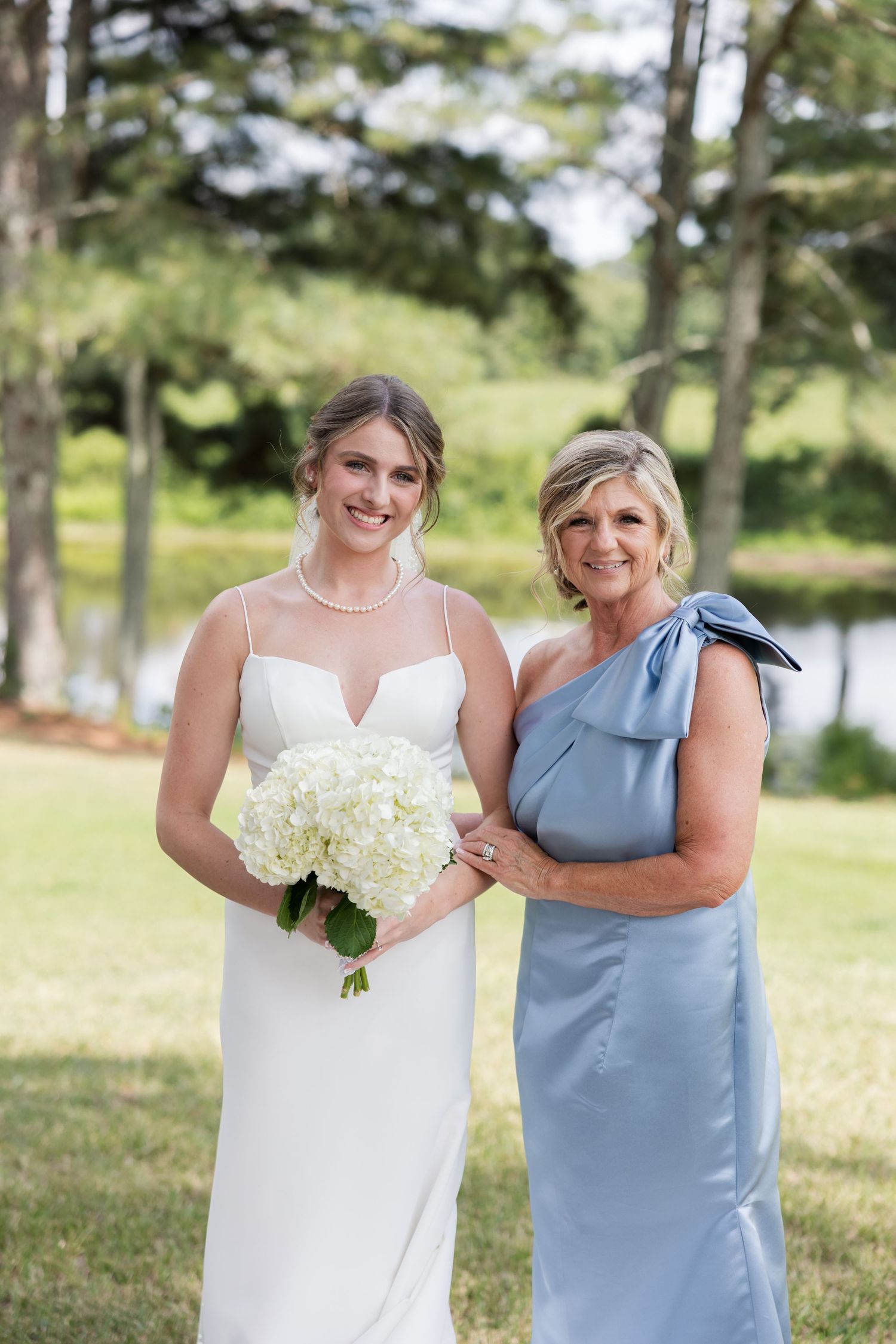 Bride in white gown holding white hydrangea bouquet poses with woman in blue one-shoulder dress outdoors.