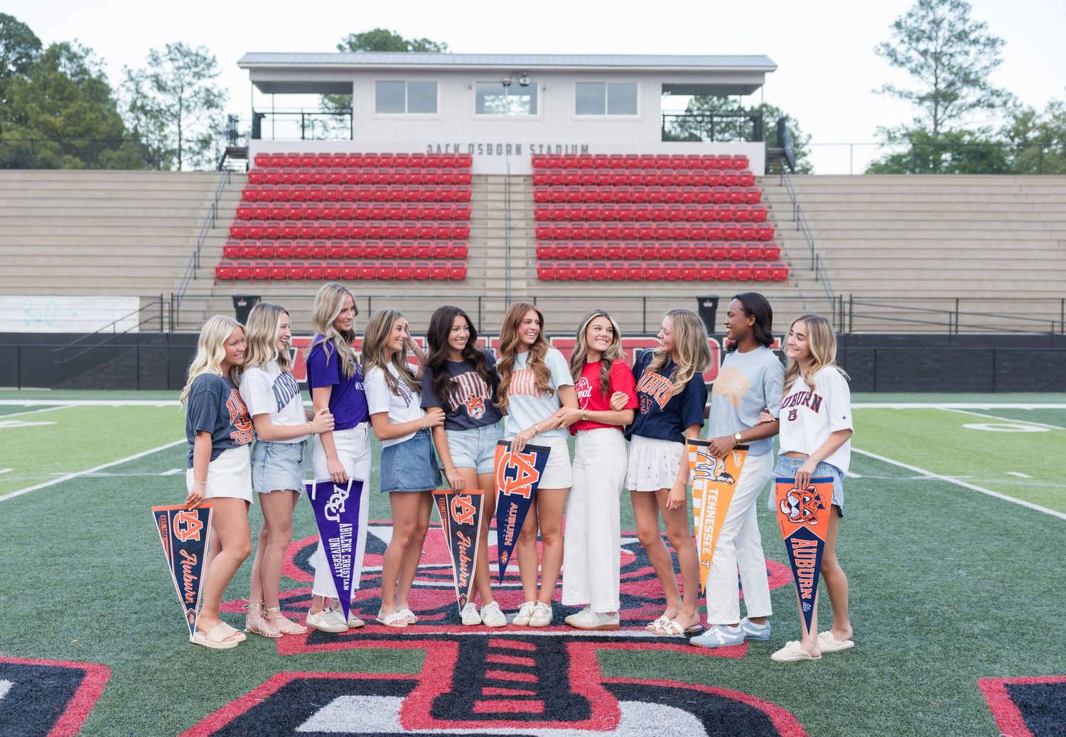 A group of teenage girls posing together on a football field holding colorful pennants, with red stadium bleachers behind them.