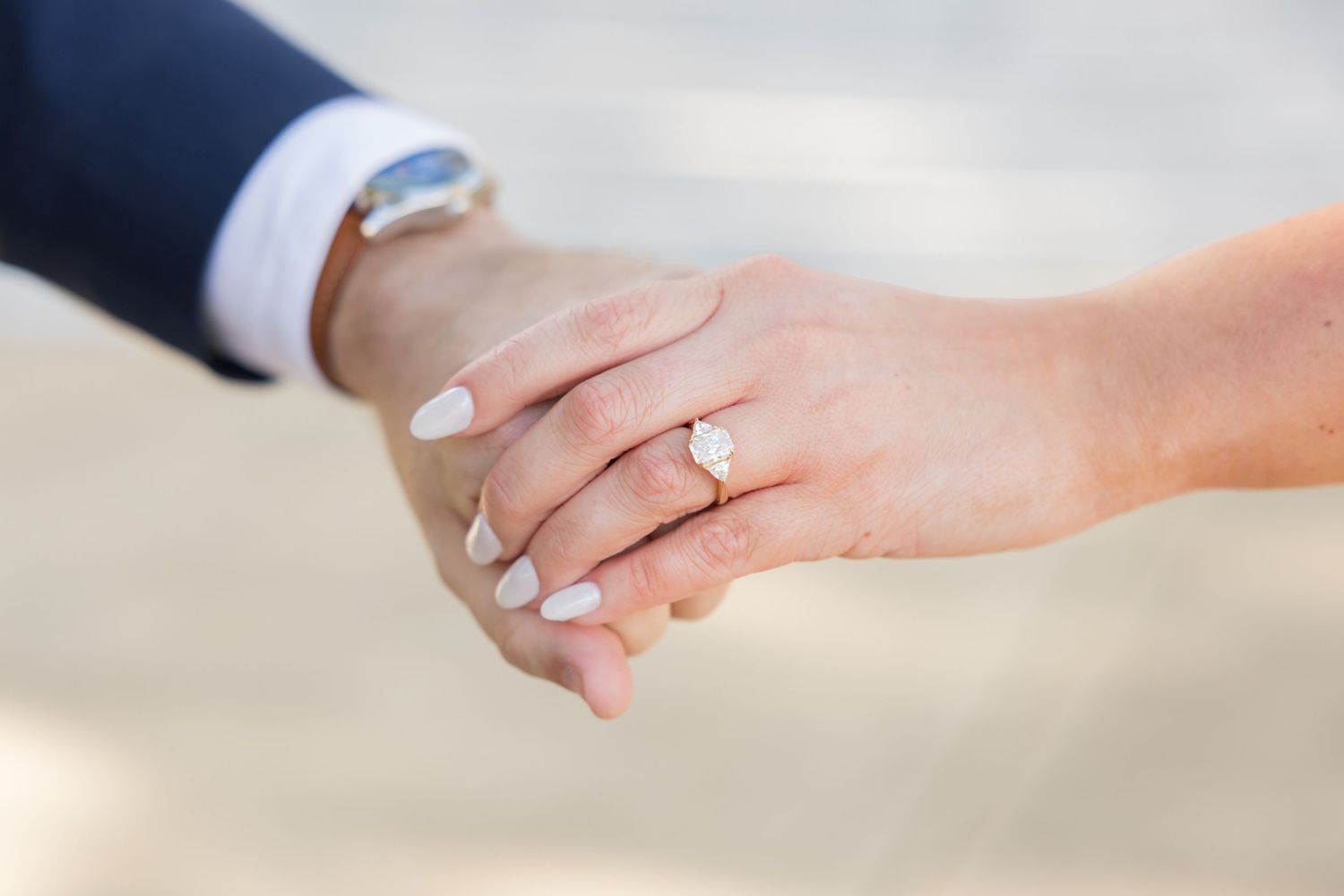 Close-up of bride and groom holding hands, showcasing diamond engagement ring with white manicured nails.