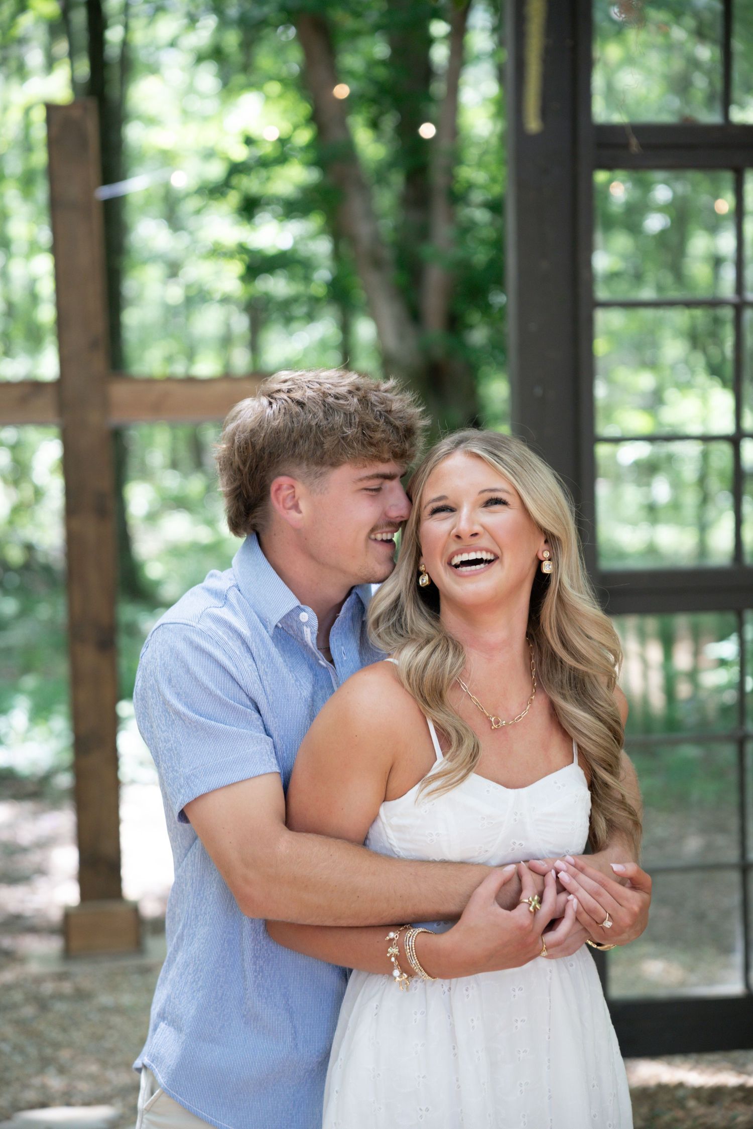 Happy couple embracing outdoors, woman in white dress laughing joyfully as man holds her from behind near rustic windows.
