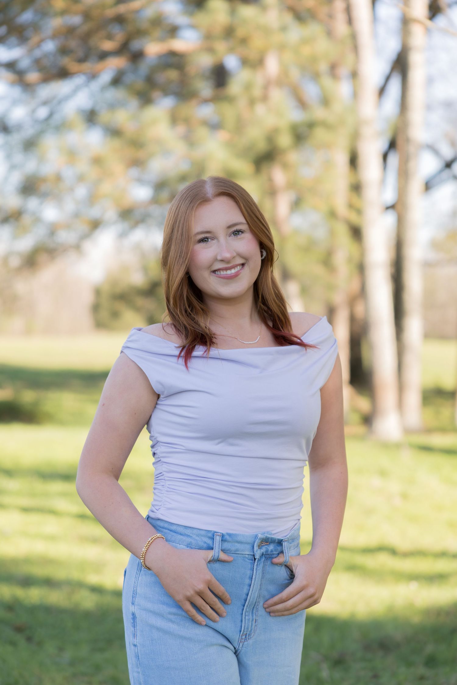 Young woman with red hair smiling outdoors in a white off-shoulder top and jeans in a sunny park.