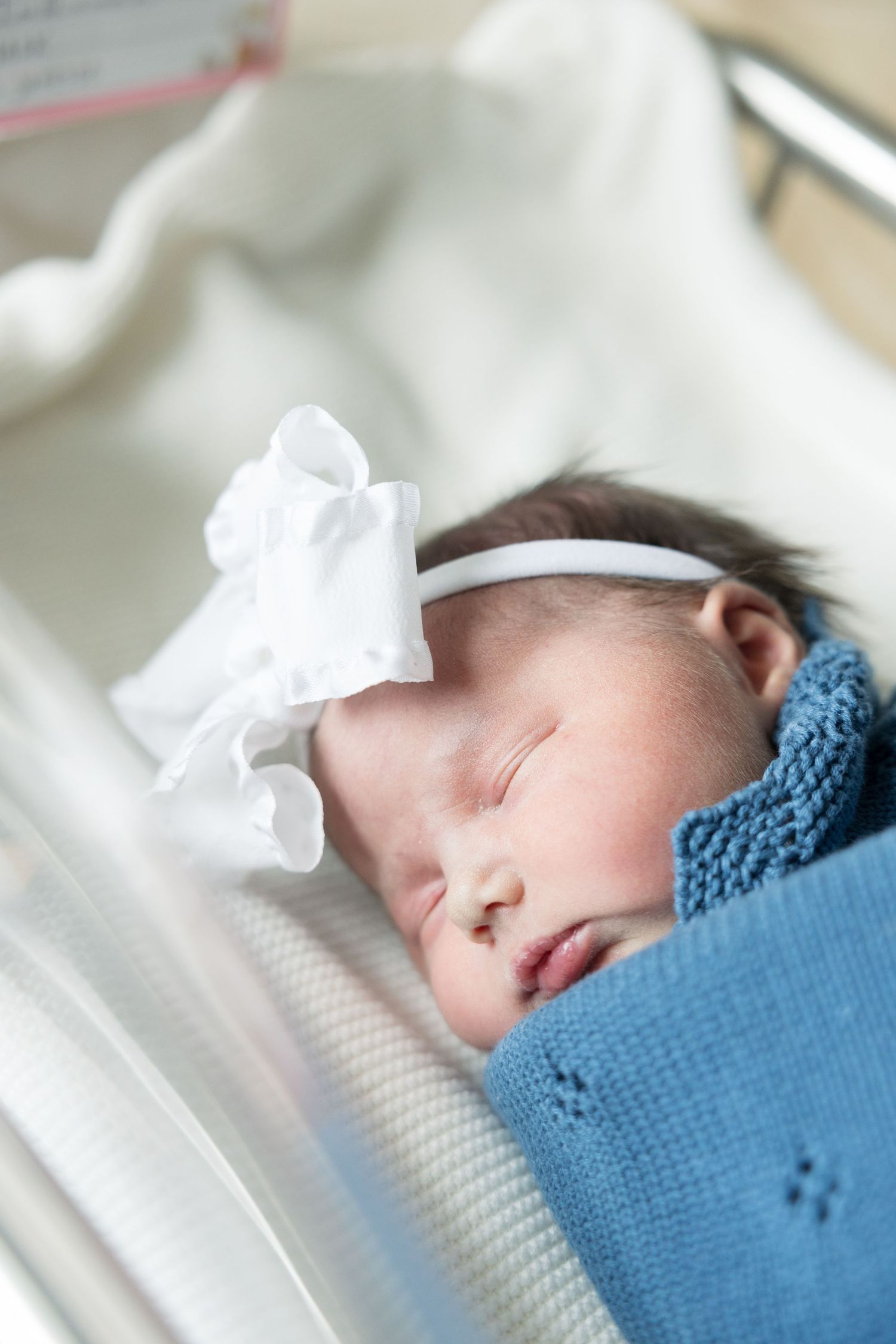 Newborn baby girl sleeping peacefully in hospital bassinet, wrapped in blue blanket with white bow headband.