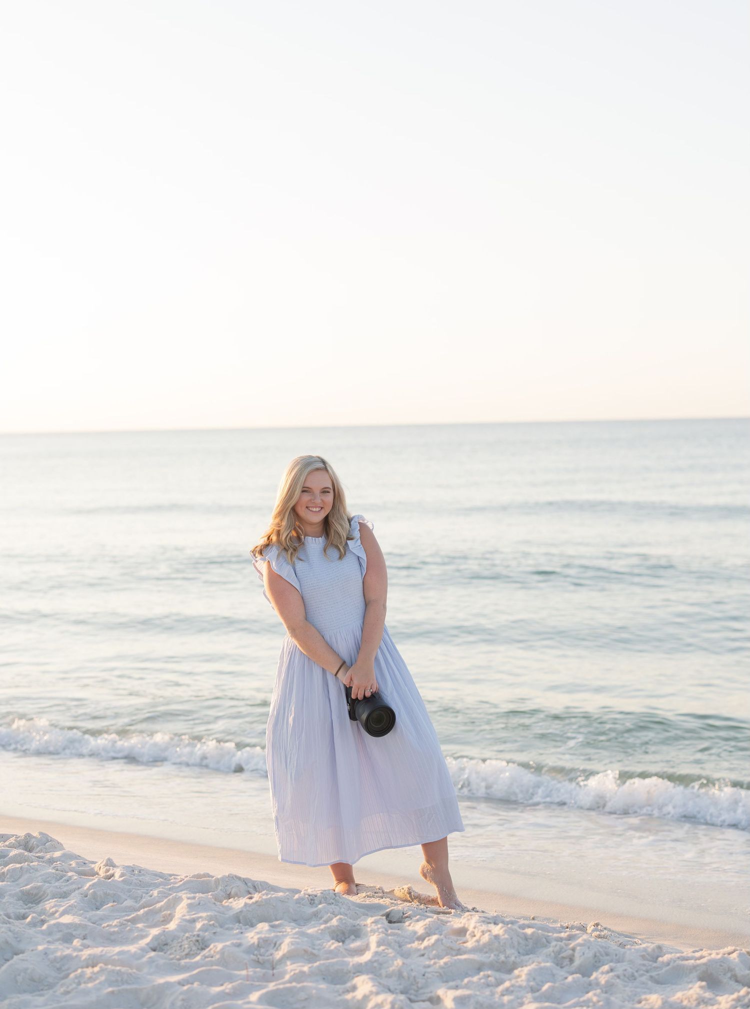 Smiling female photographer in light blue midi dress holds a camera lens on a bright sandy beach.