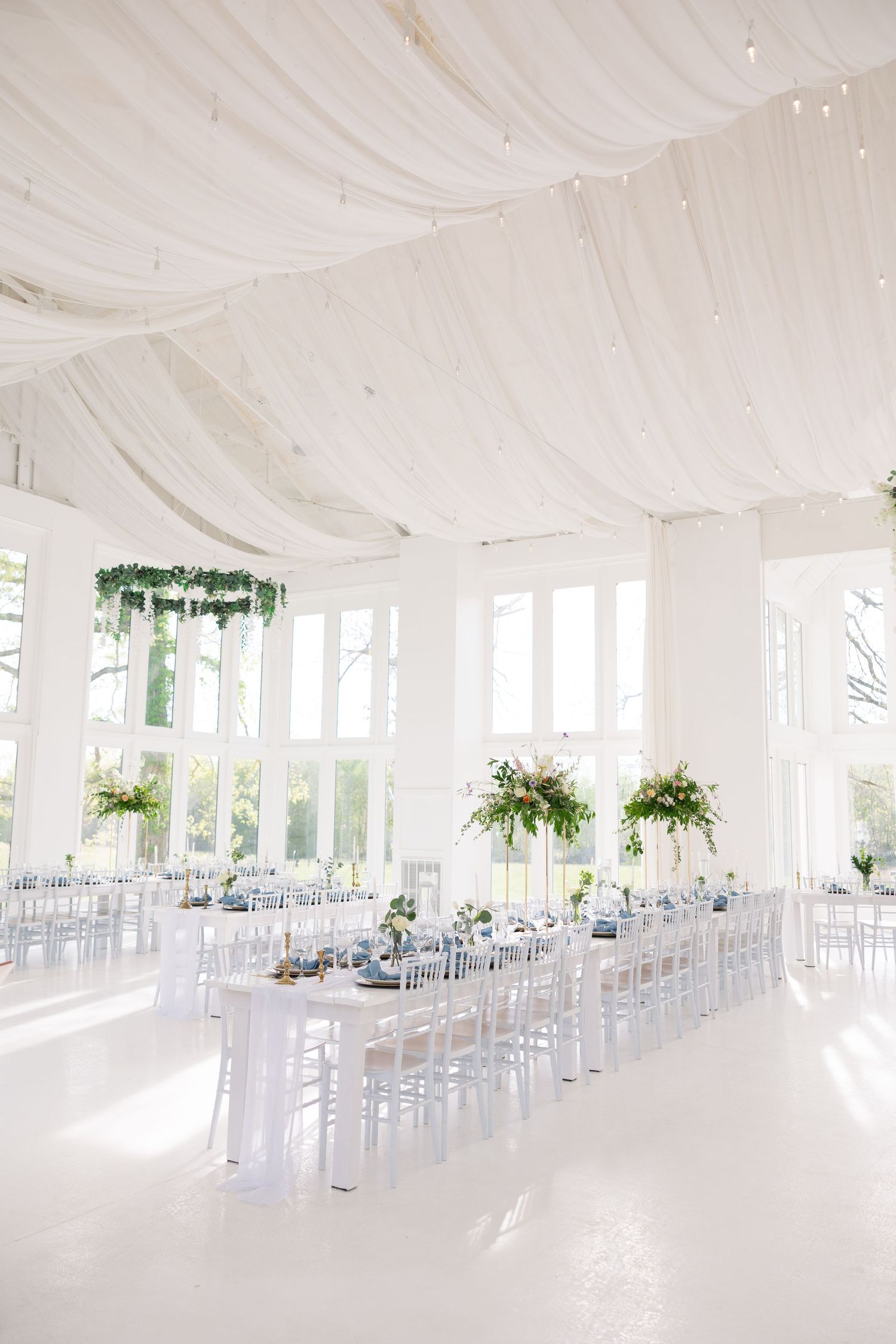 Elegant white wedding reception hall with draped ceiling, long tables, ghost chairs, and lush greenery floral arrangements.