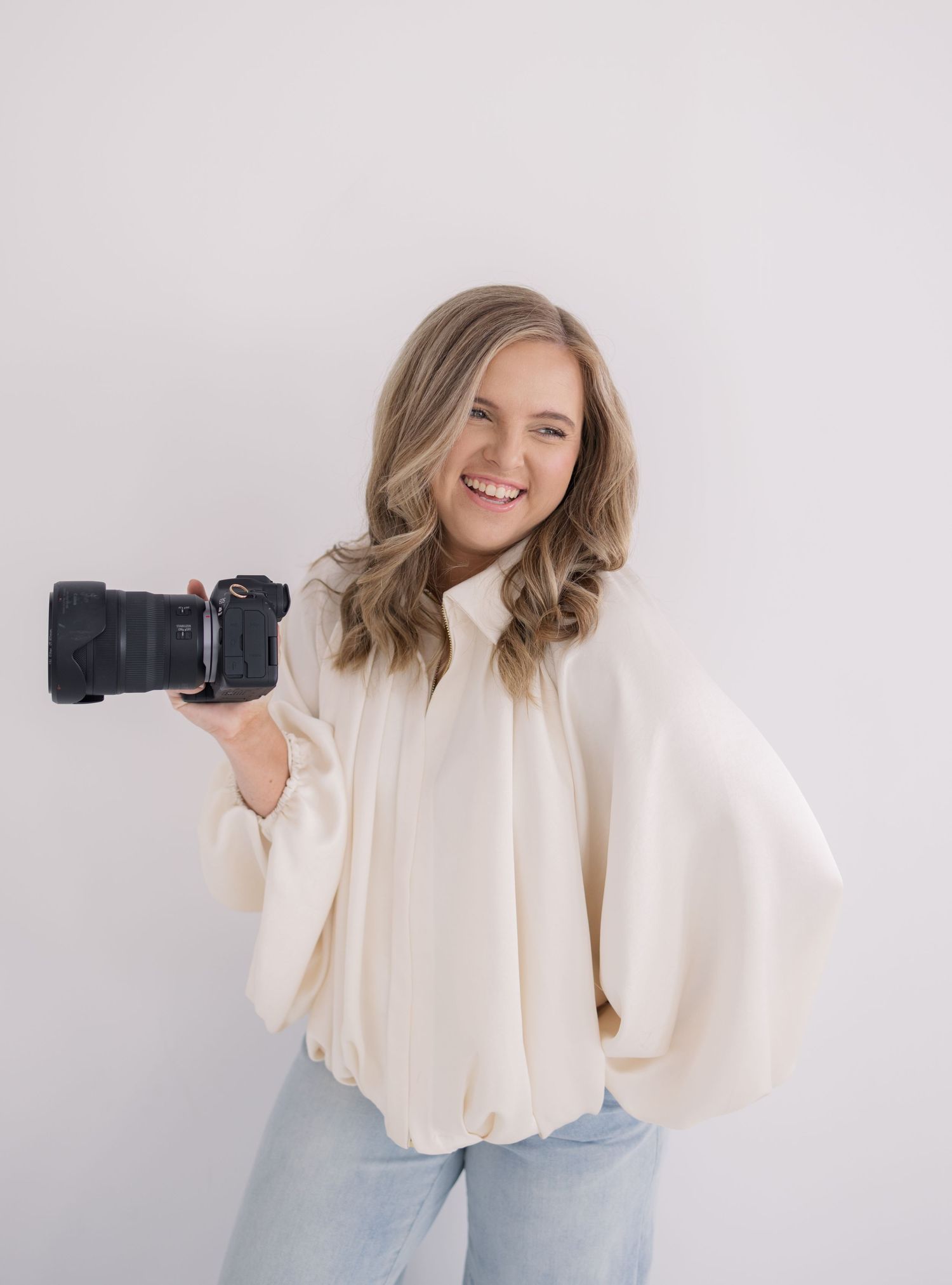 Smiling woman photographer holding a DSLR camera, wearing cream blouse and jeans, posing against white background.